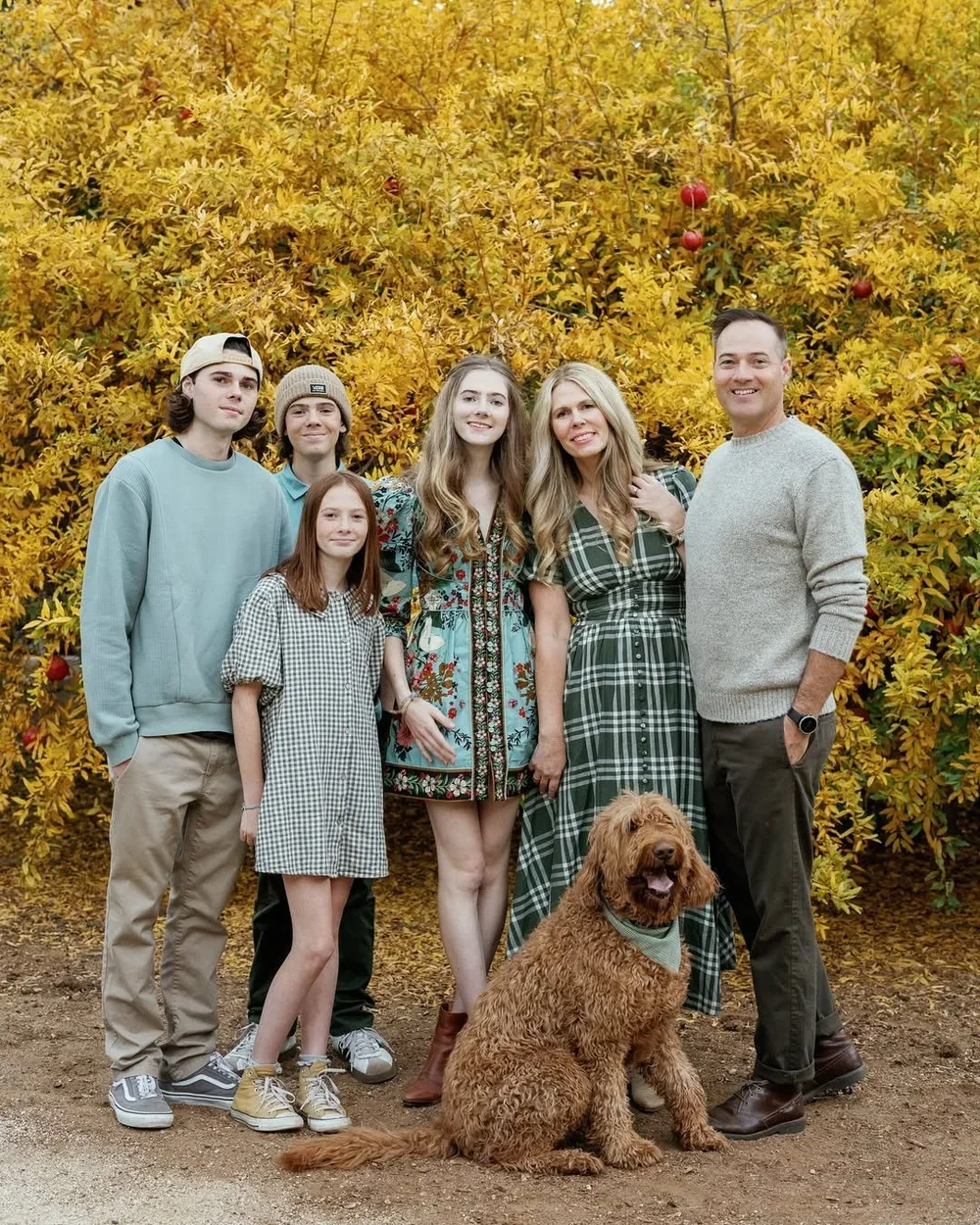 A family of six people and a dog posing outdoors in front of a yellow leafy bush with red apples. The group includes three women, two men, and two girls, all smiling and dressed casually.