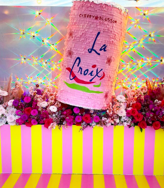 A pink cotton candy machine with a sign that reads "La Croix" and the flavor "Cherry Blossom," surrounded by pink, red, and white flowers, and a background with rainbow-colored star-shaped light reflections, all set against a yellow and pink striped base.