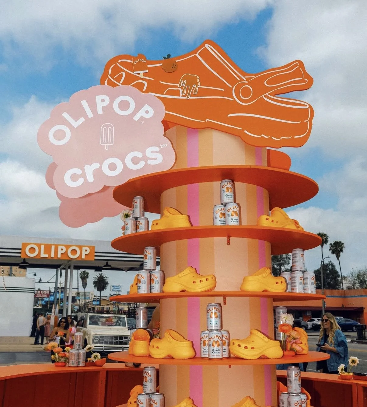 A colorful outdoor display featuring a large pink and orange structure resembling a multi-tiered cake with Croc shoes and cans of orange cream soda. The sign reads 'OLIPOP crocs,' with a large cartoon croc outlined in orange on top. People are walking in the background near a store called OLIPOP.