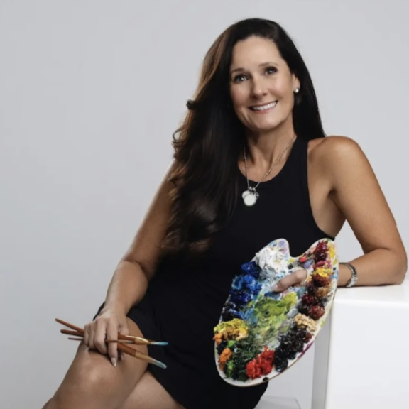 A woman with long dark hair, wearing a black sleeveless dress, holding paintbrushes and a colorful painter's palette. She is smiling and posing against a plain white background.