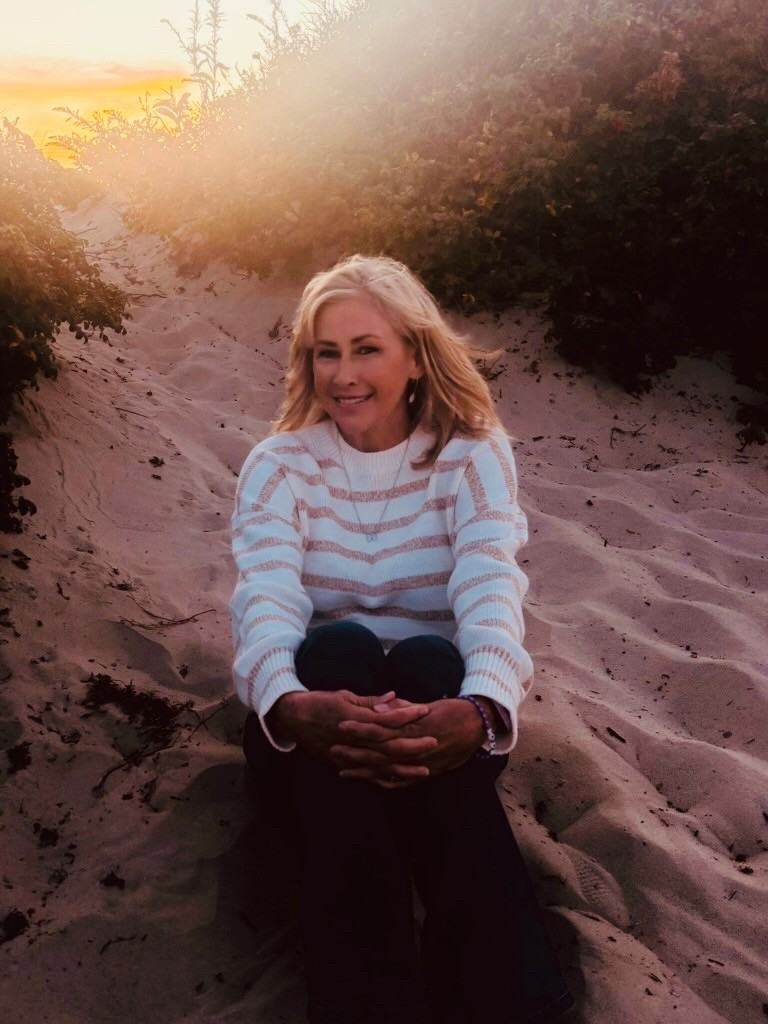 A woman with blonde hair sitting on sand with her knees drawn to her chest, smiling at the camera, with bushes and sunset in the background.