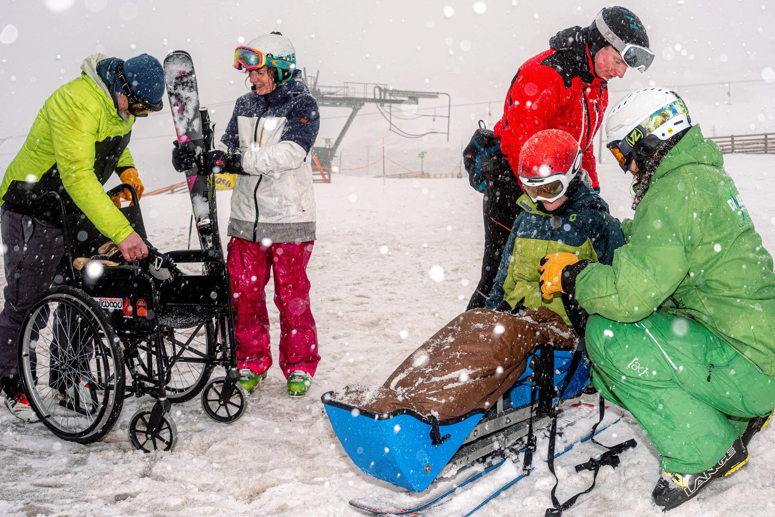 Helpers prepare a young skier in a blue sit-ski beside a wheelchair as snow falls.