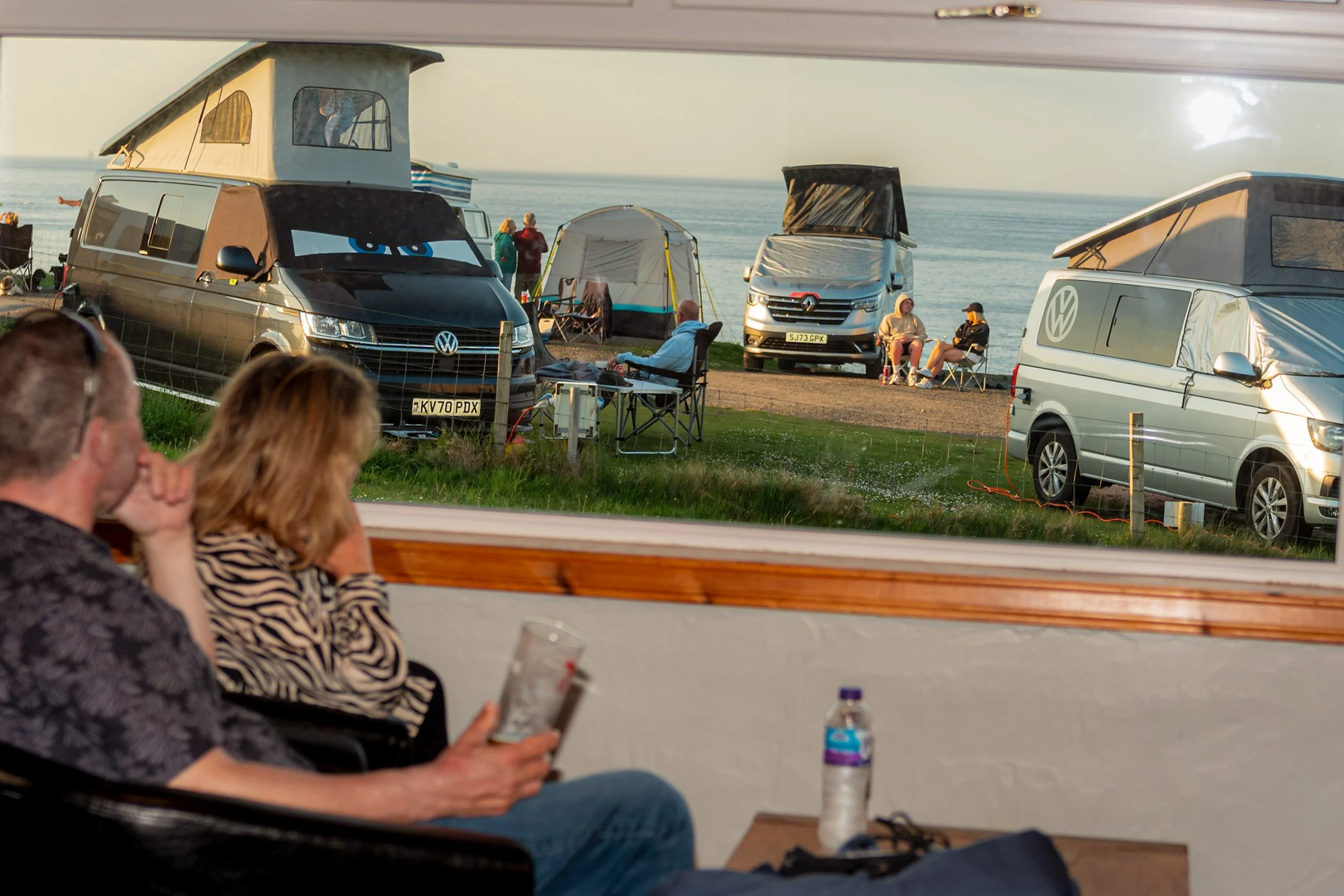 A couple look out of a window at camper vans parked along a grassy clifftop.