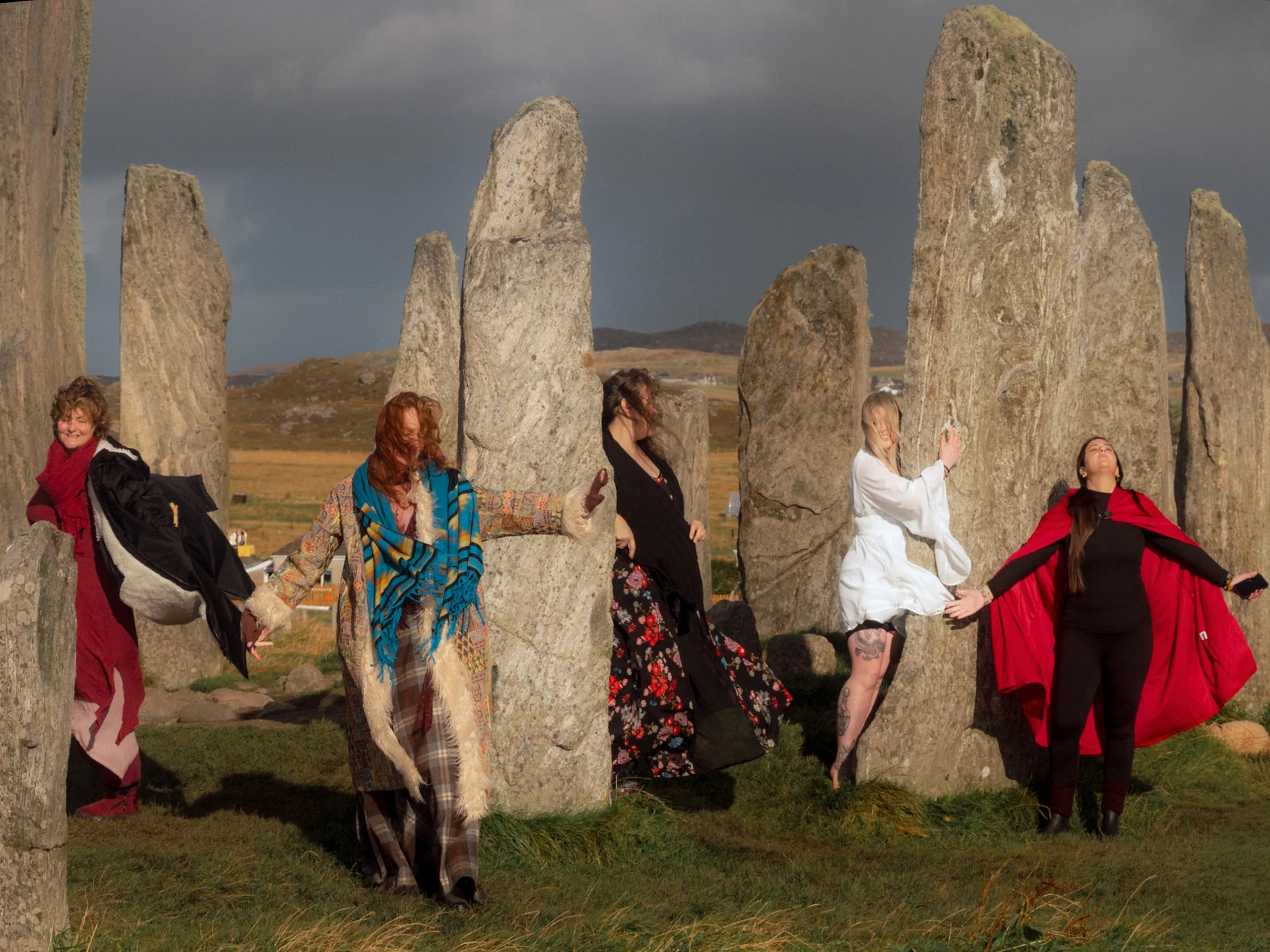 Women in long cloaks and dresses pose among tall standing stones.