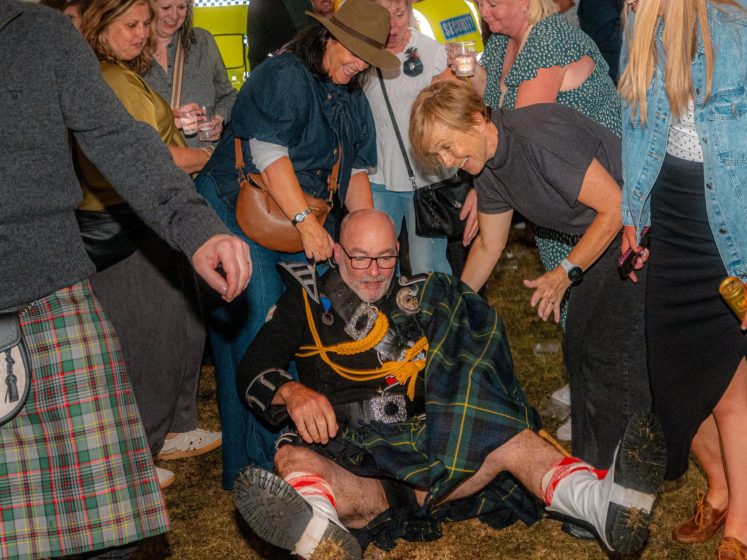 A man in Highland dress sits on the grass as a crowd gathers round him at a Highland games.