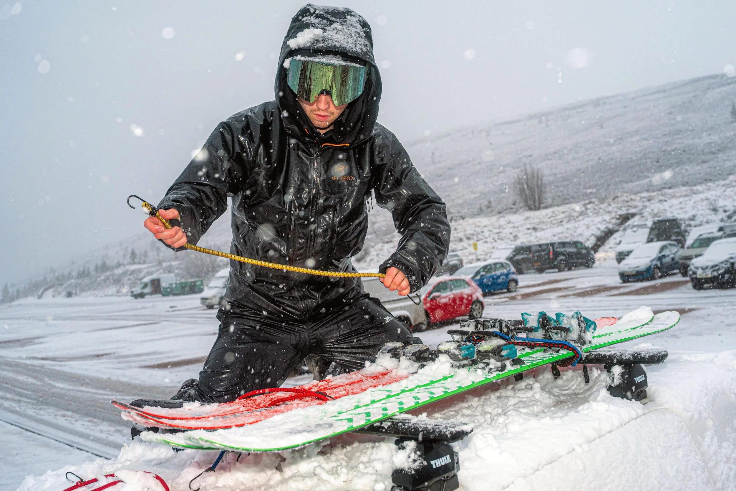 A skier in a black jacket and goggles straps his skis together in a snowy car park.