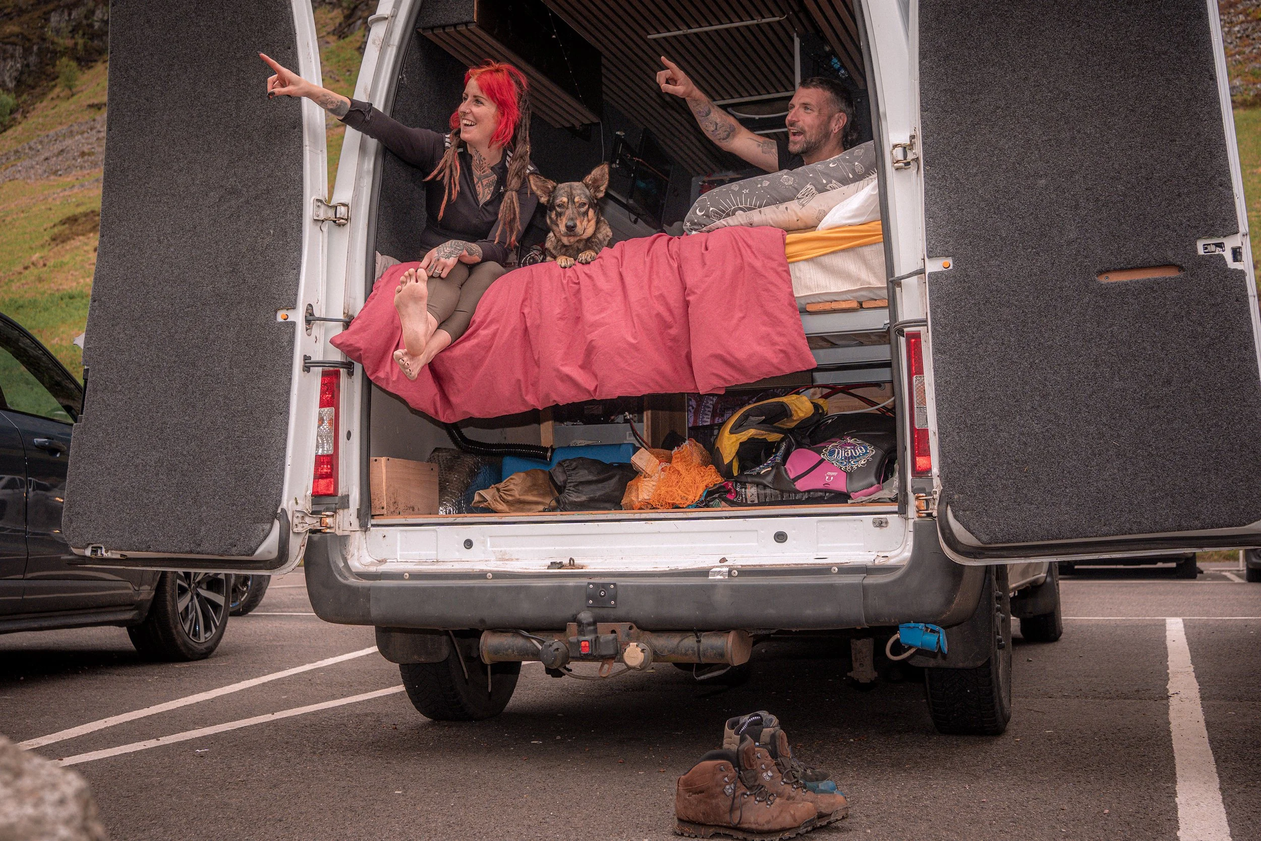 A couple and their dog relax in the back of a camper van with its doors open in a car park.