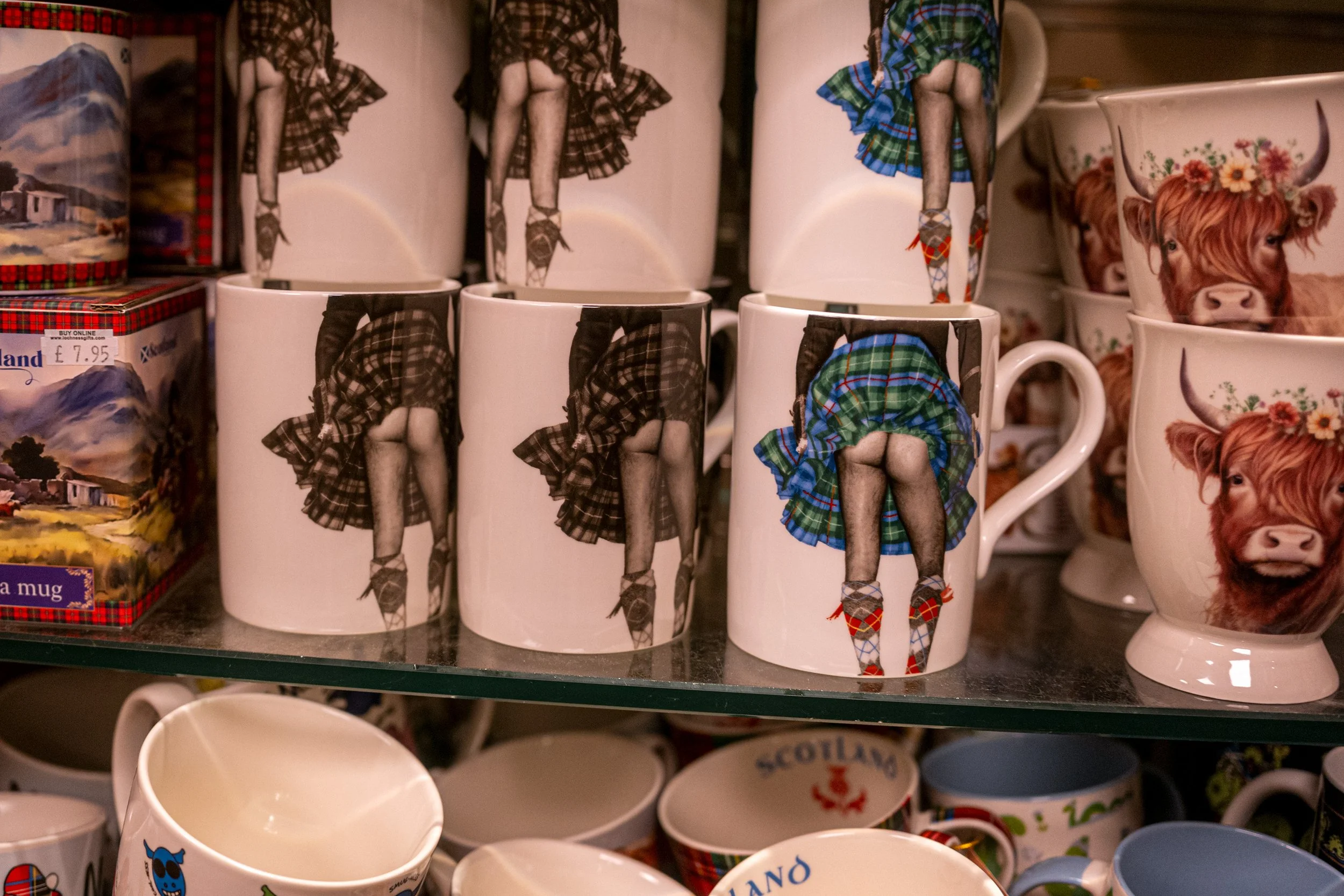 Souvenir mugs printed with kilted legs and Highland cows on a gift shop shelf.