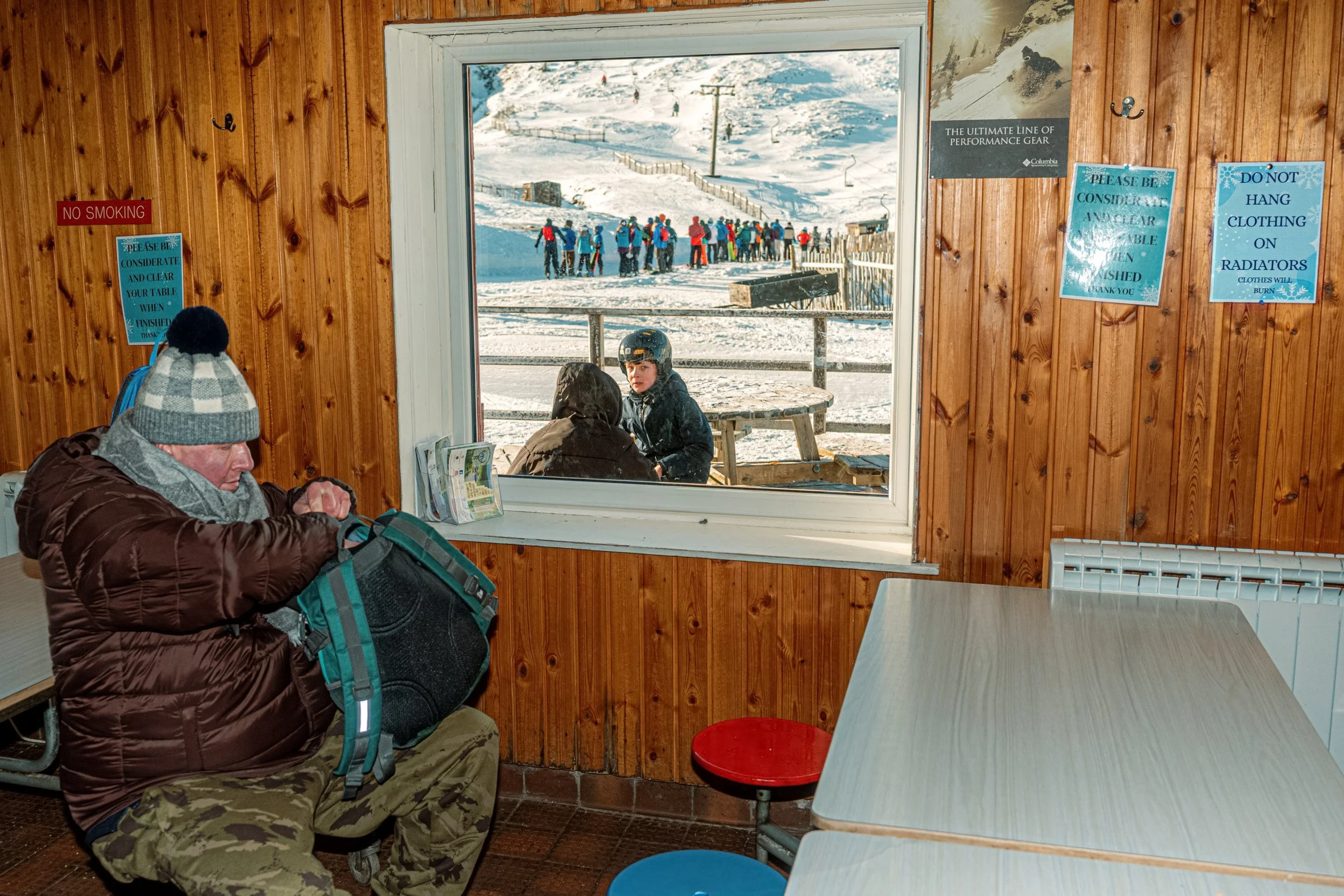 A visitor unpacks a bag inside a wooden ski-resort shelter, with skiers visible through the window, from Weather Permitting.
