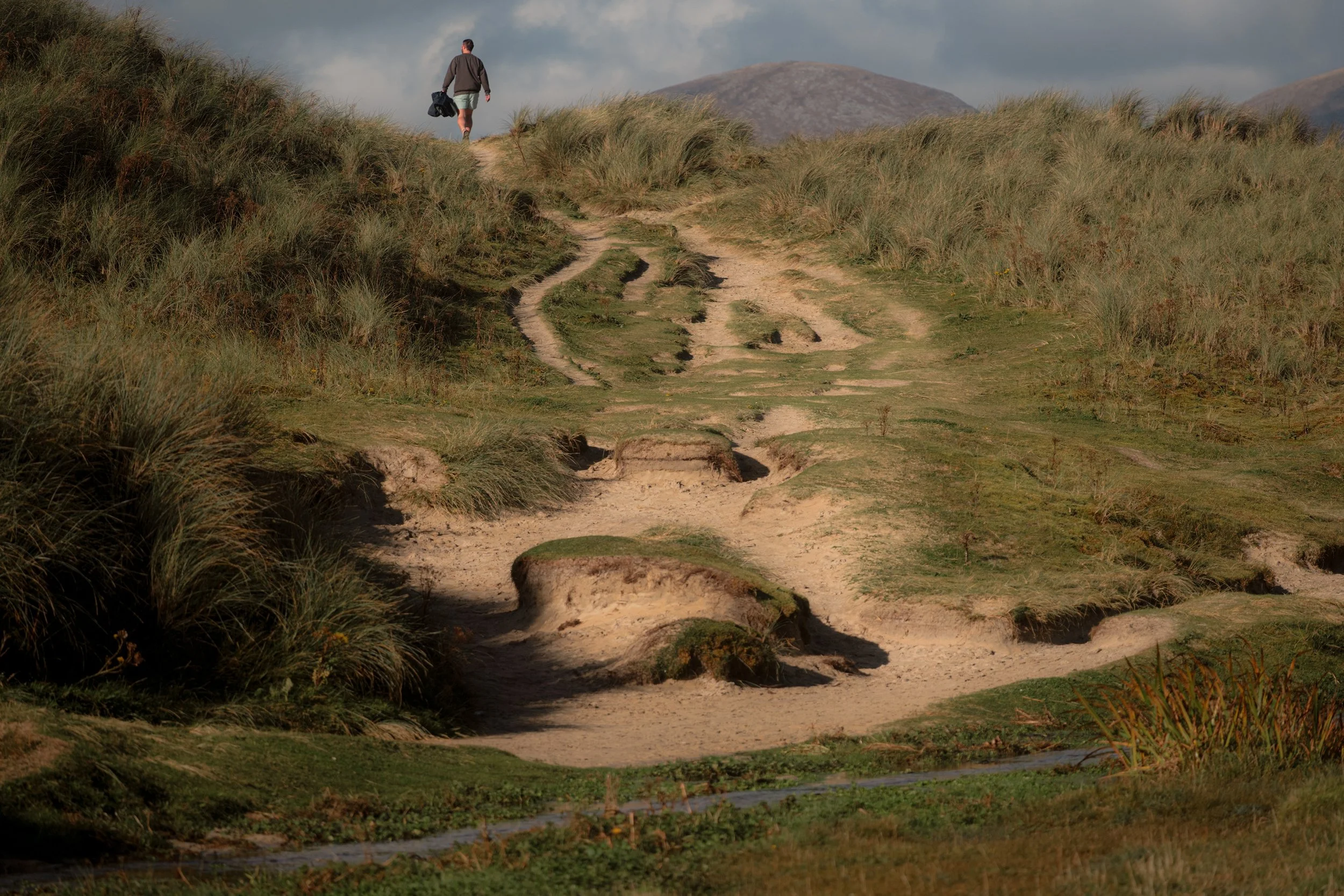 A lone walker crosses worn sand dunes on a coastal path.
