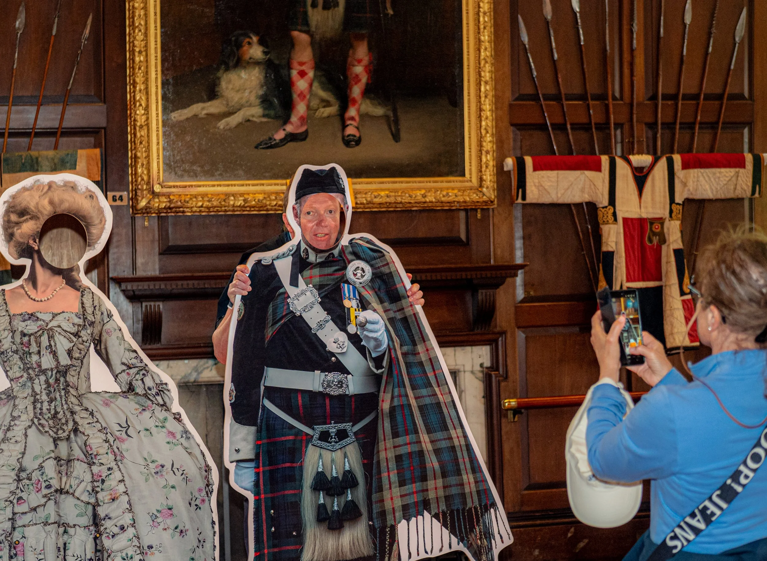 A visitor photographs life-size cut-outs of figures in Highland dress and a ball gown in a castle room.
