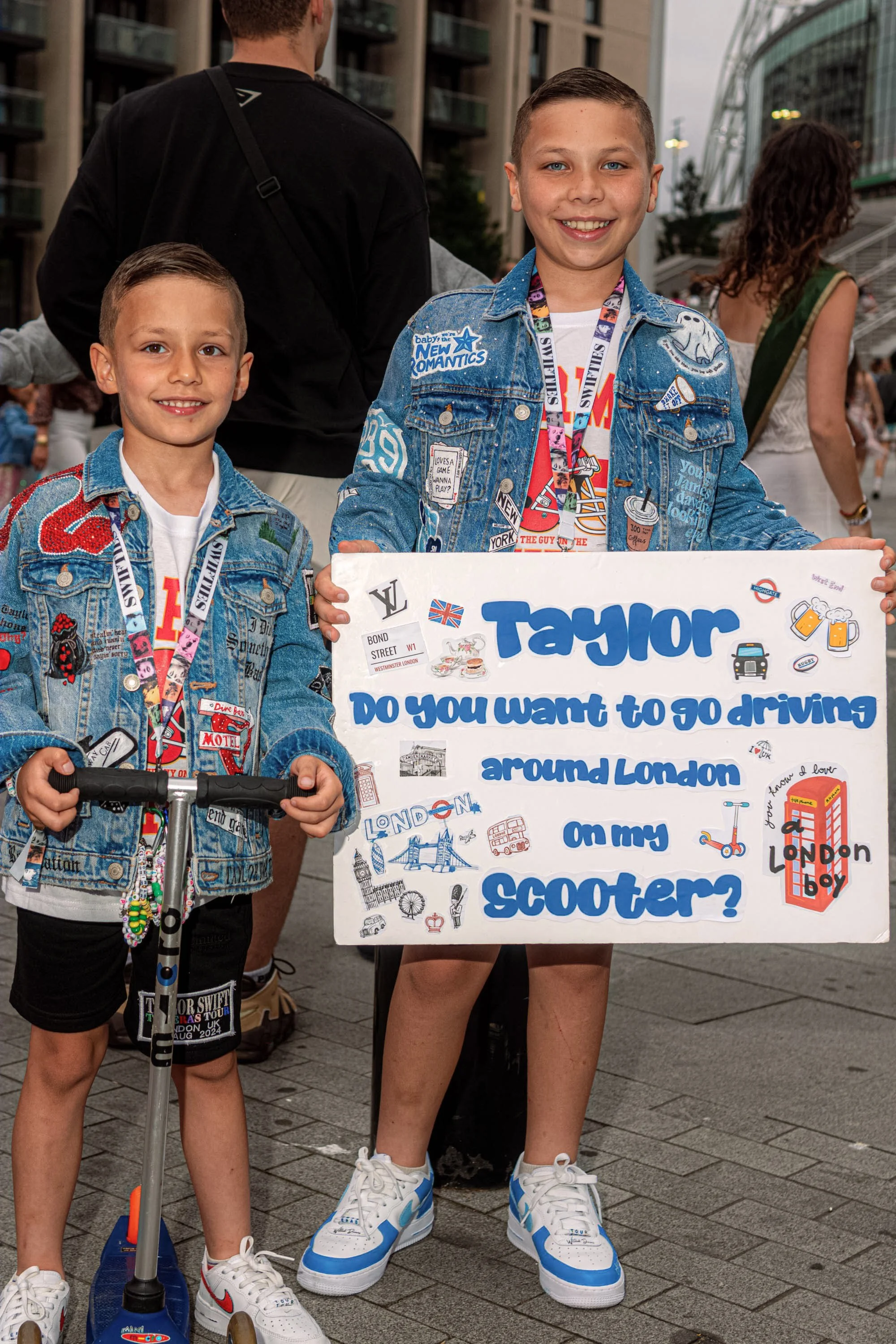 Two young Taylor Swift fans in decorated denim jackets hold a sign inviting Taylor to ride scooters around London.