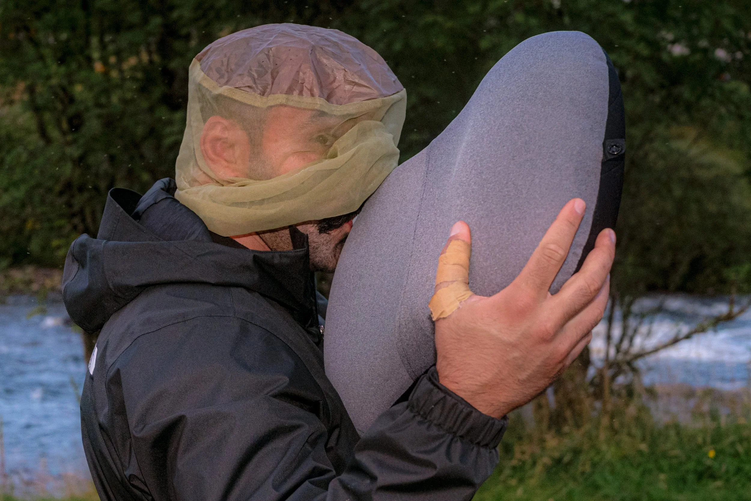 A man in a midge net carries a large grey bag beside a river.