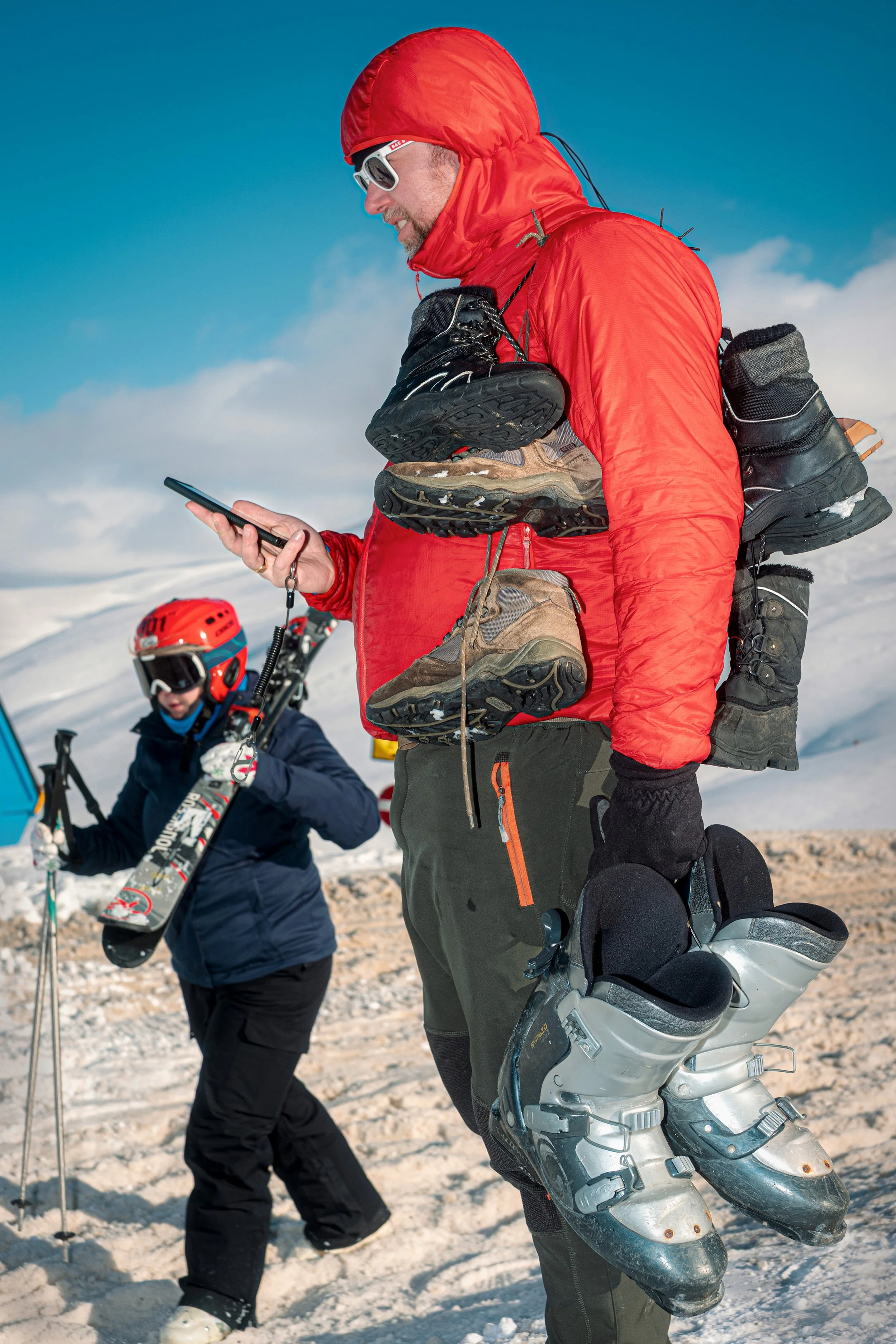 A man with ski boots hanging from his rucksack checks his phone beside a child carrying skis.
