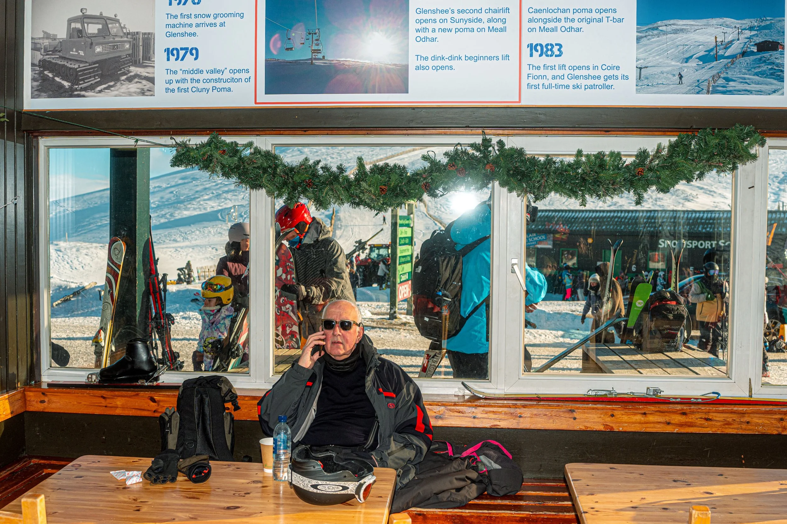 A skier on the phone sits at a café table beneath a ski history display, with a Christmas garland over the window.