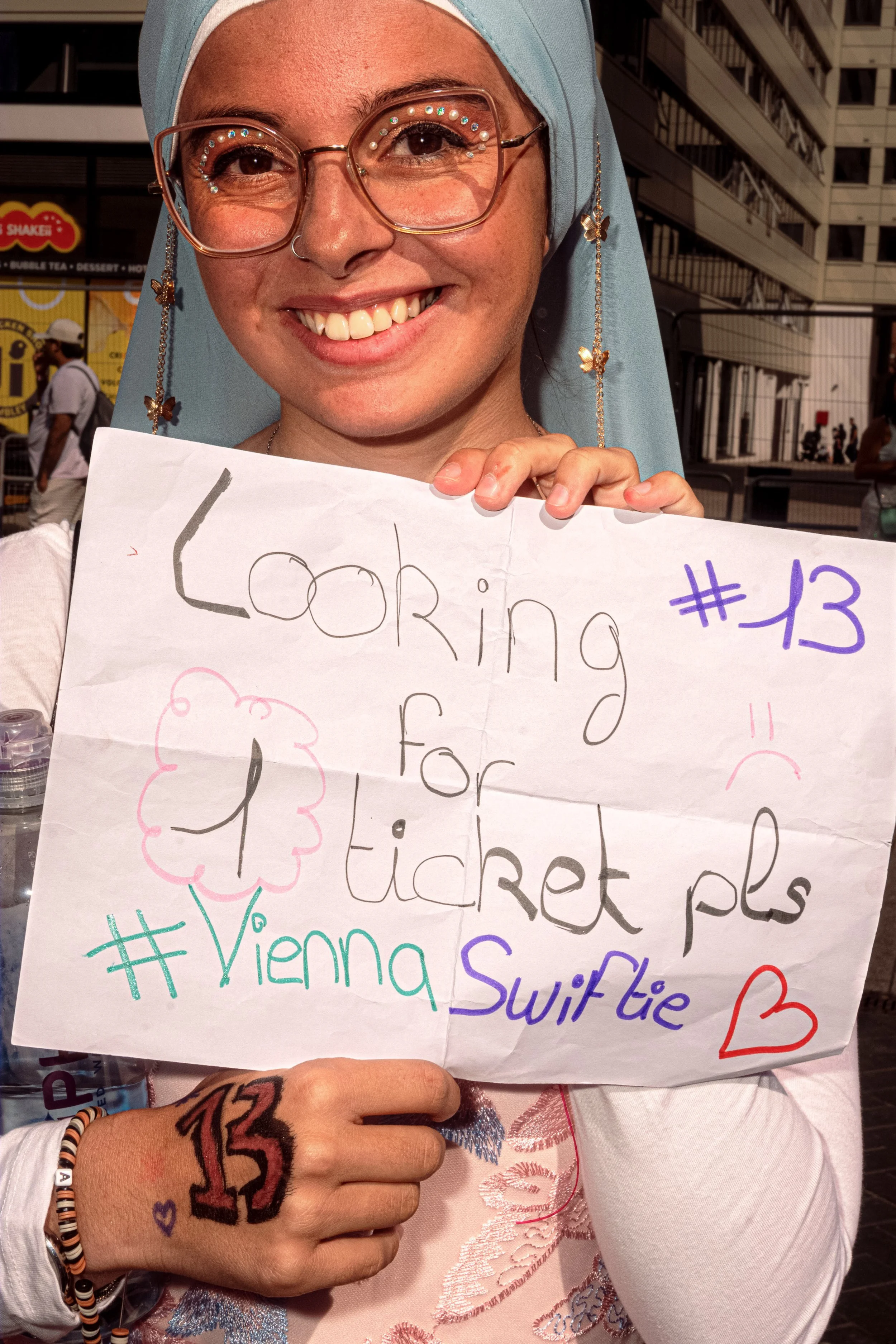 A smiling Taylor Swift fan holds a handmade sign asking for a ticket, with the number 13 drawn on her hand.