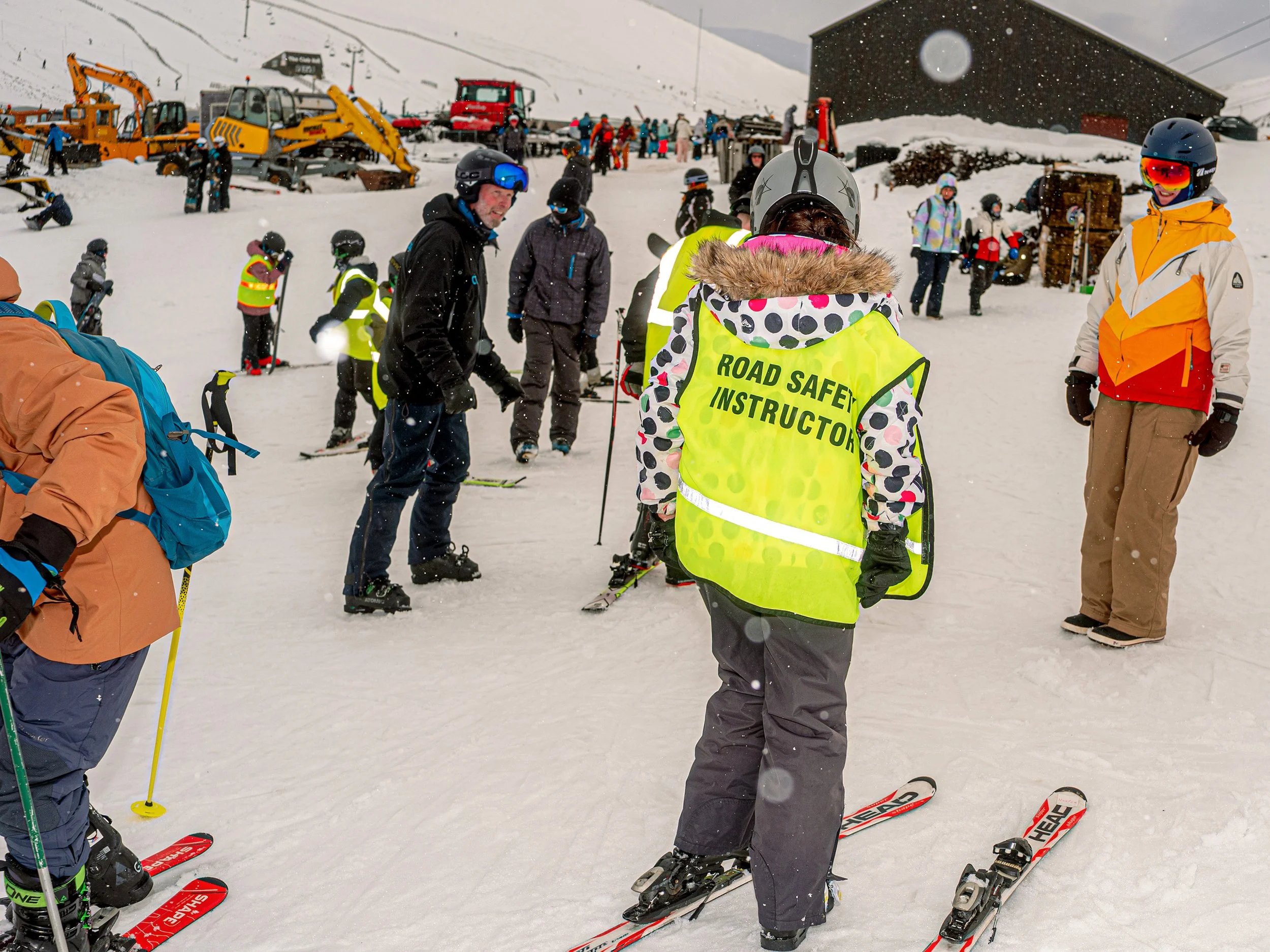 A road safety instructor in a hi-vis tabard stands among skiers and diggers at a busy ski centre.