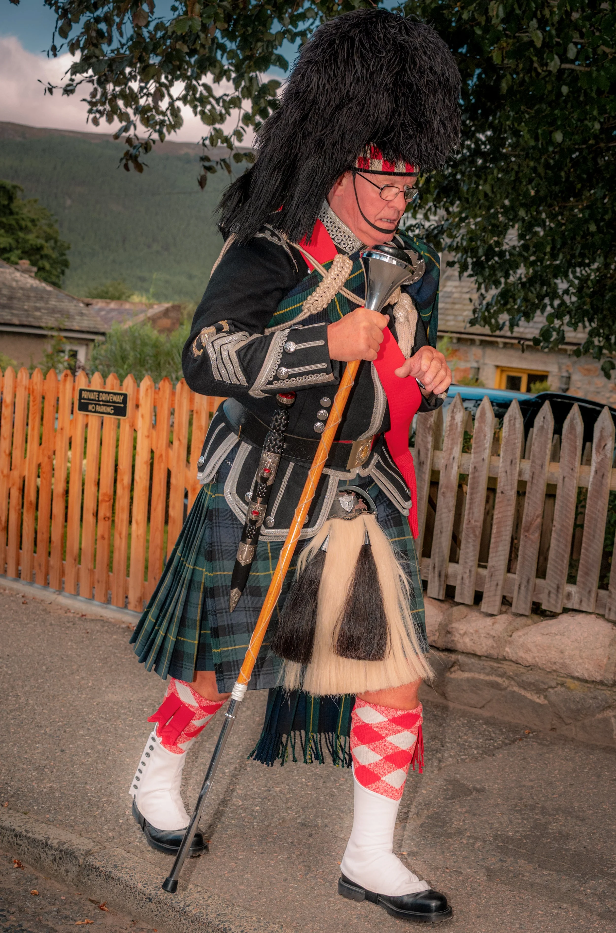 A piper in full Highland dress and feather bonnet walks along a pavement.