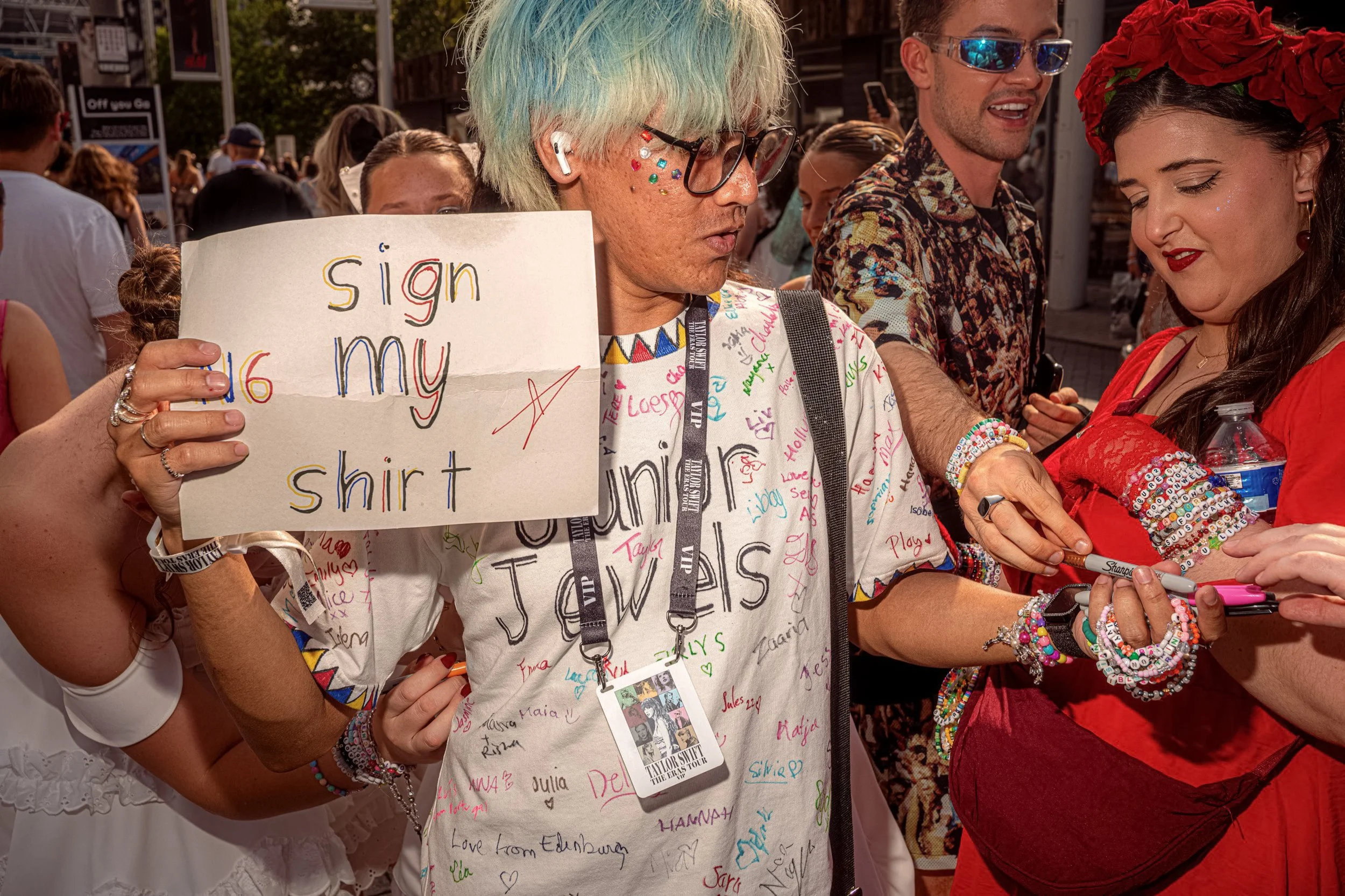 A Taylor Swift fan with pale blue hair holds a “Sign my shirt” sign while another fan writes on the T-shirt.