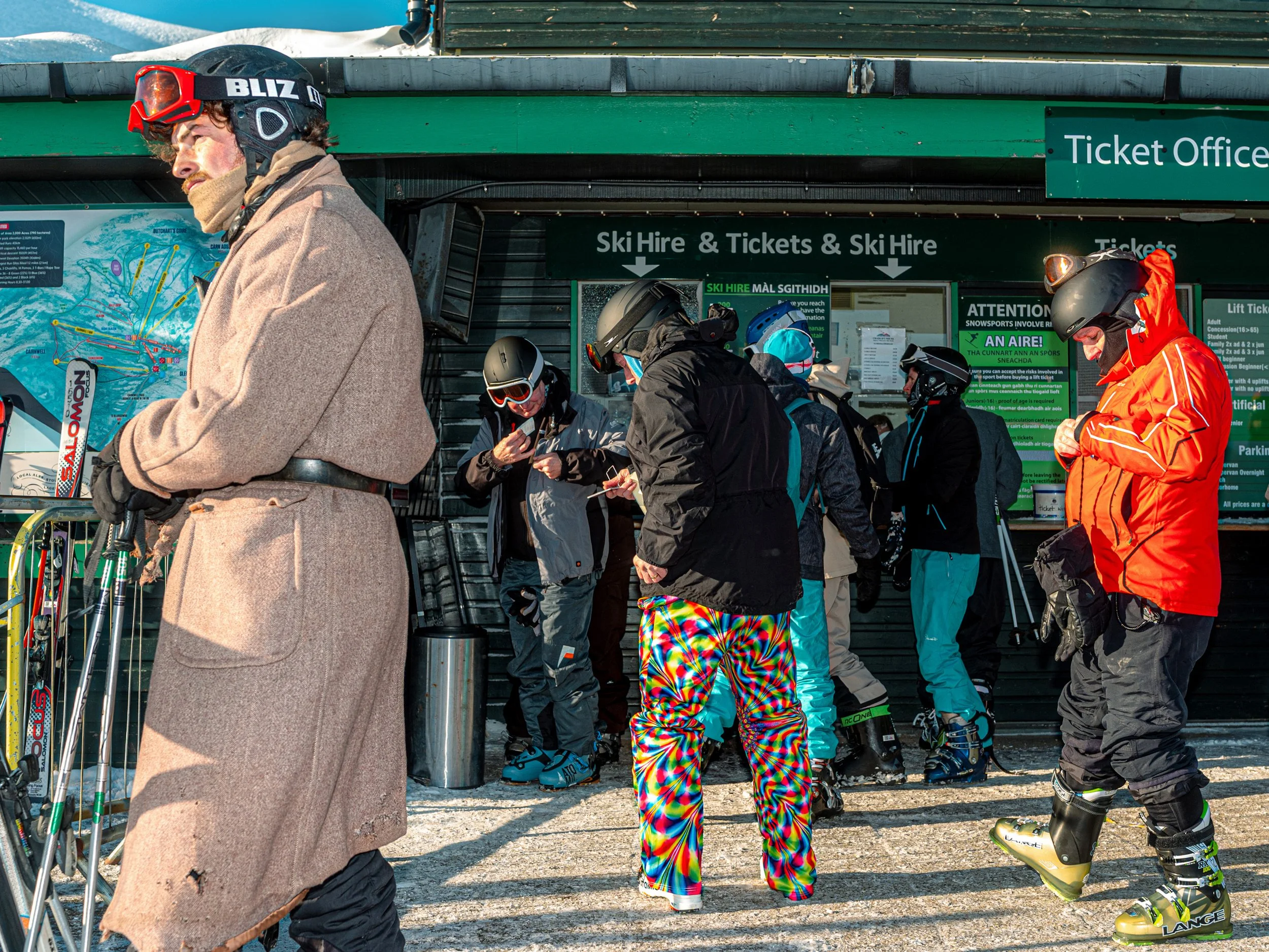 A skier in a long tweed coat and goggles stands outside the ski hire and ticket office among other skiers.