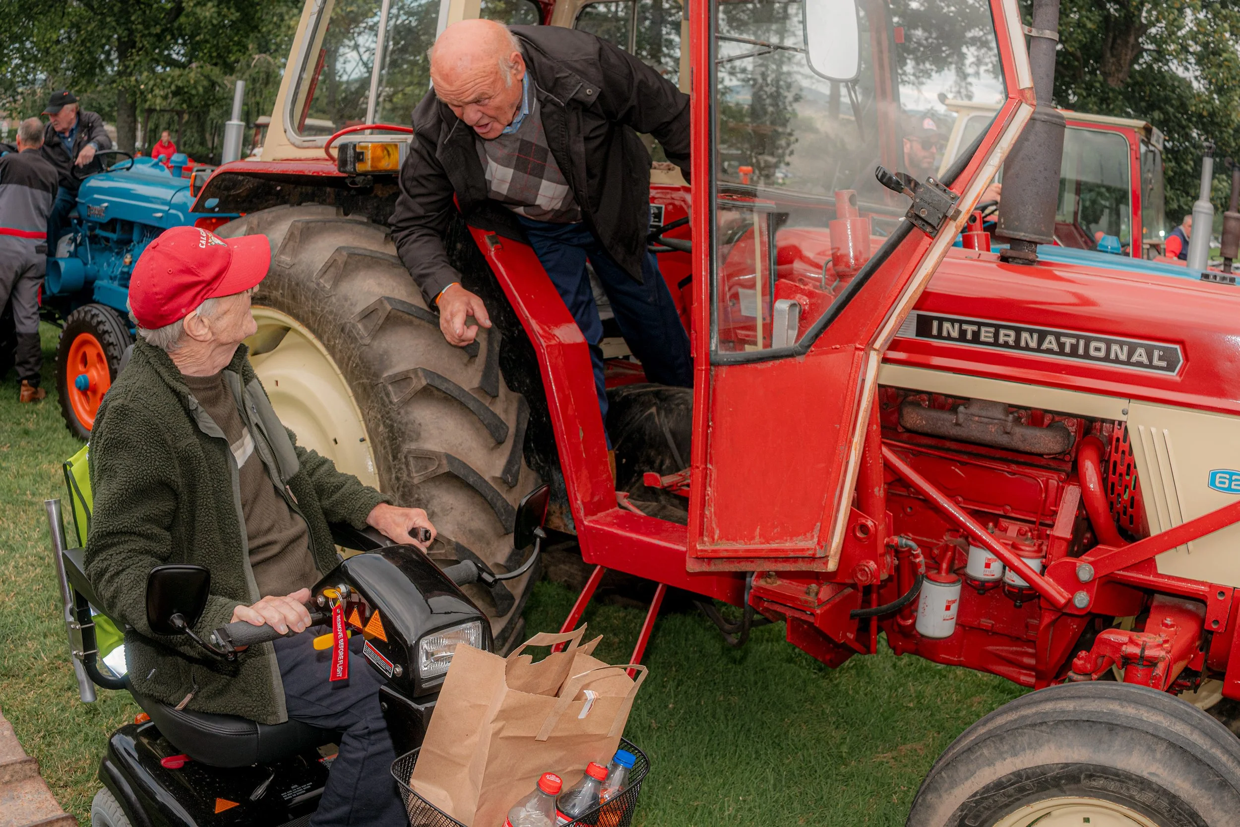 An older man climbs down from a red International tractor as a woman in a red cap sits on a mobility scooter.
