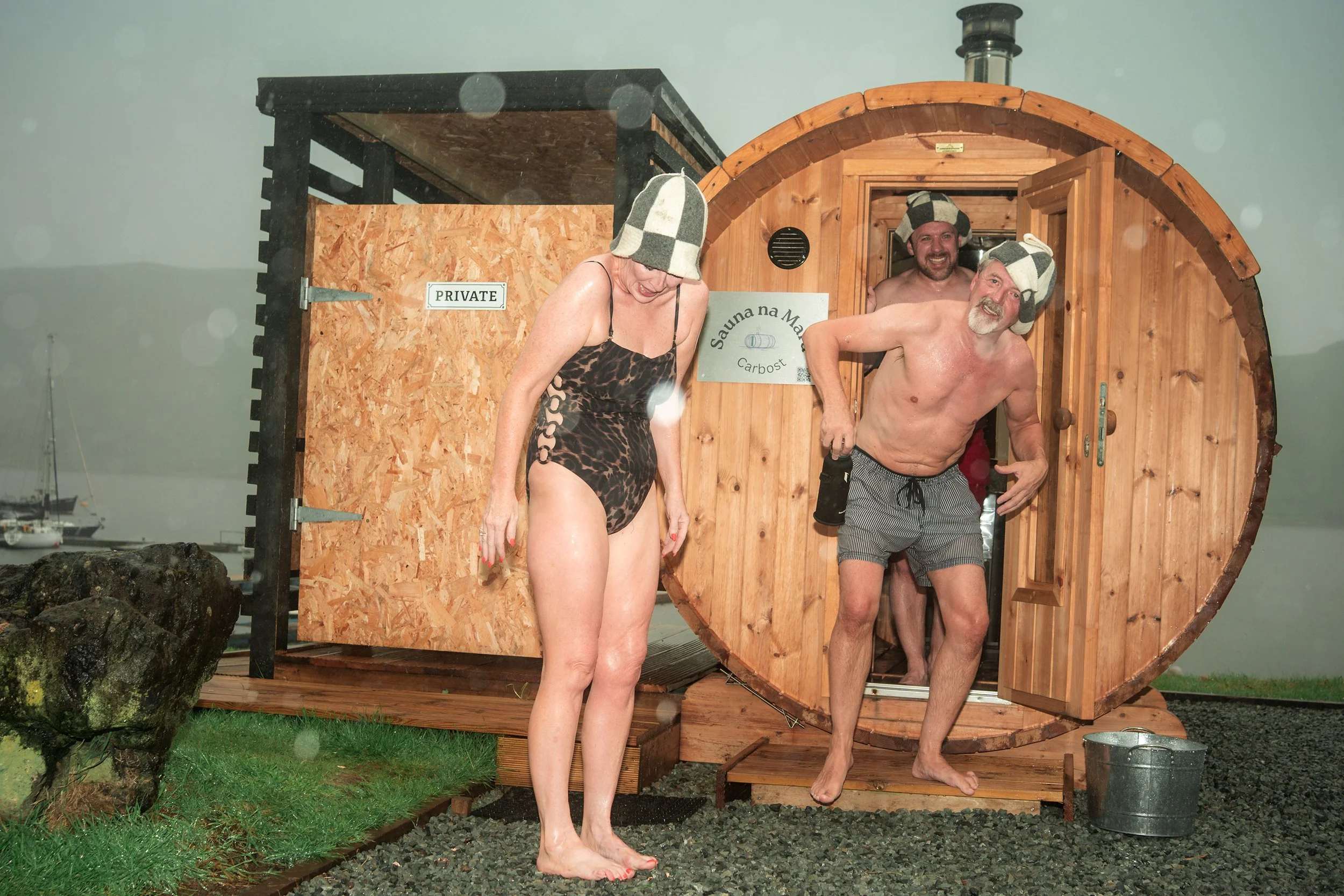 A woman and a man in felt sauna hats step out of a wooden barrel sauna by the water.