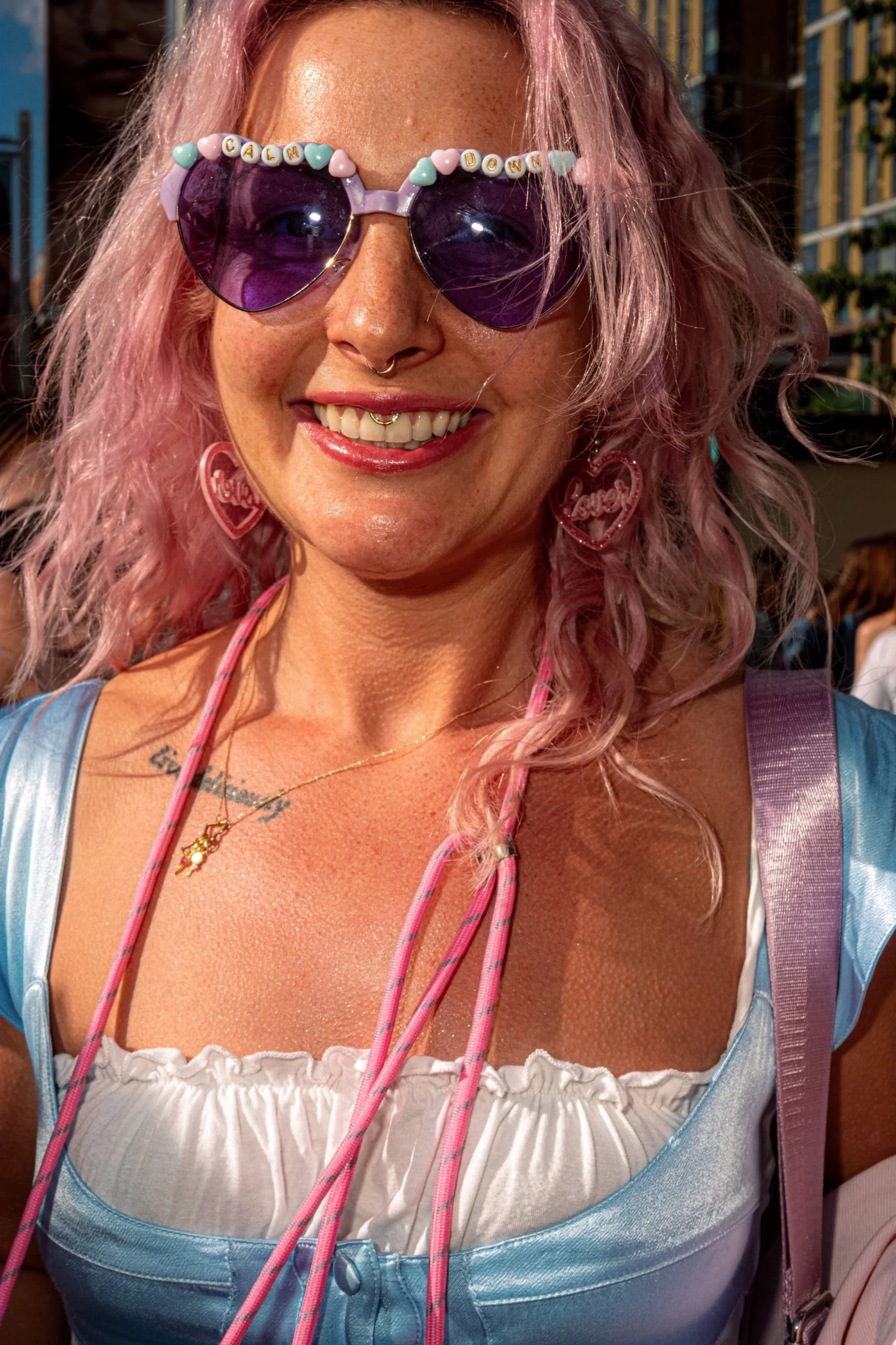 A smiling Taylor Swift fan with pink hair wears bead-trimmed heart-shaped sunglasses and a blue-and-white outfit.