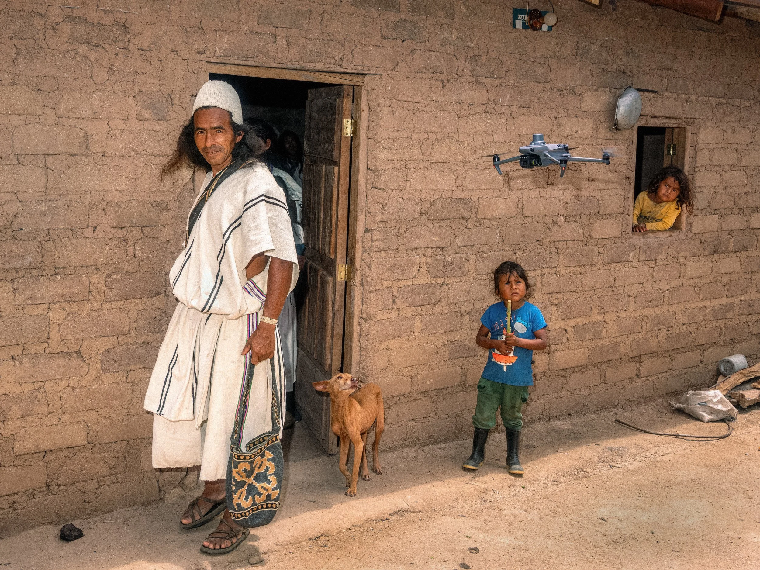 People and a dog stand outside an earth-brick house while a small drone hovers nearby.