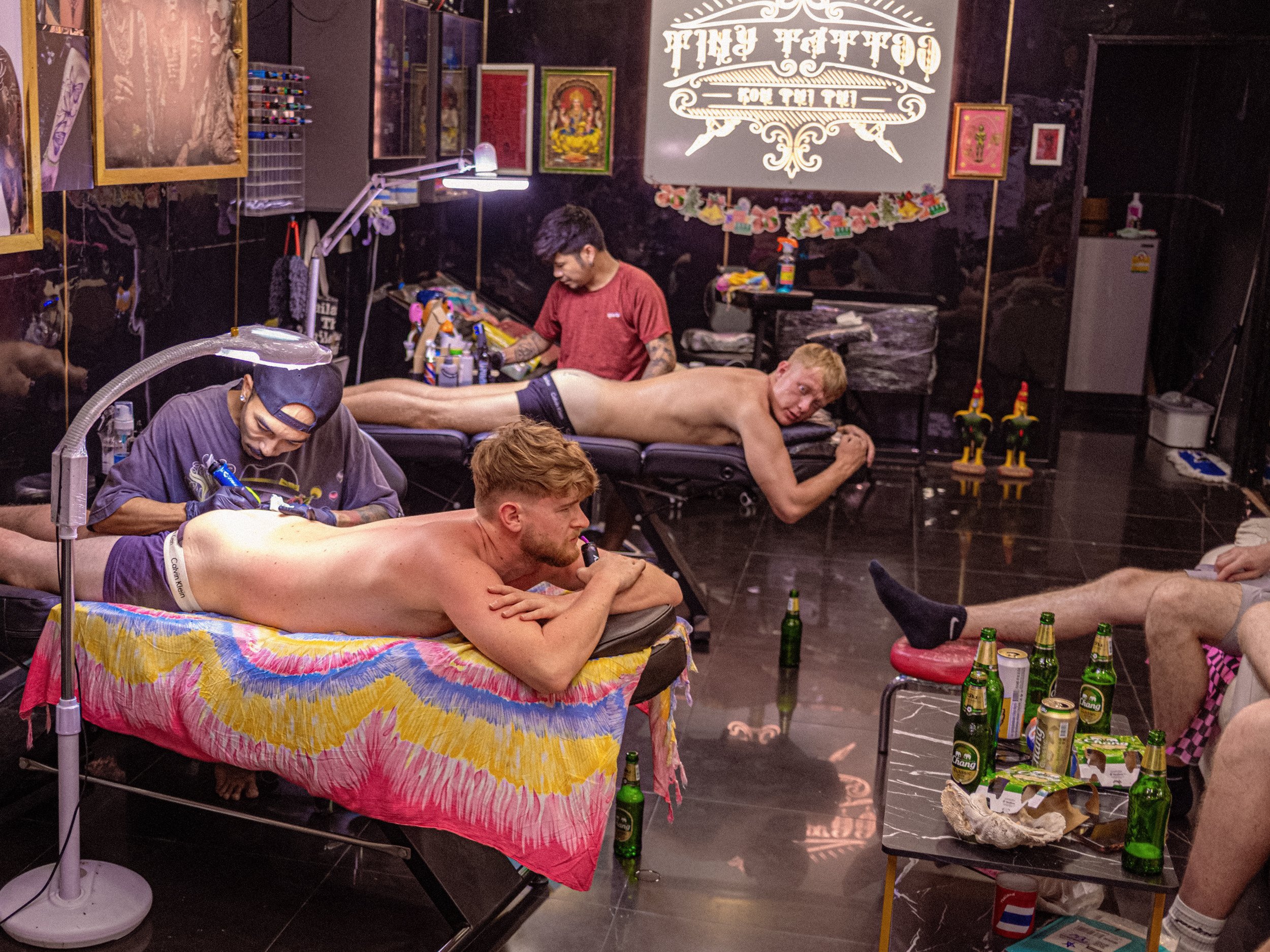 Customers receive tattoos in a tattoo studio in Thailand, with bottles arranged beside the workstations.