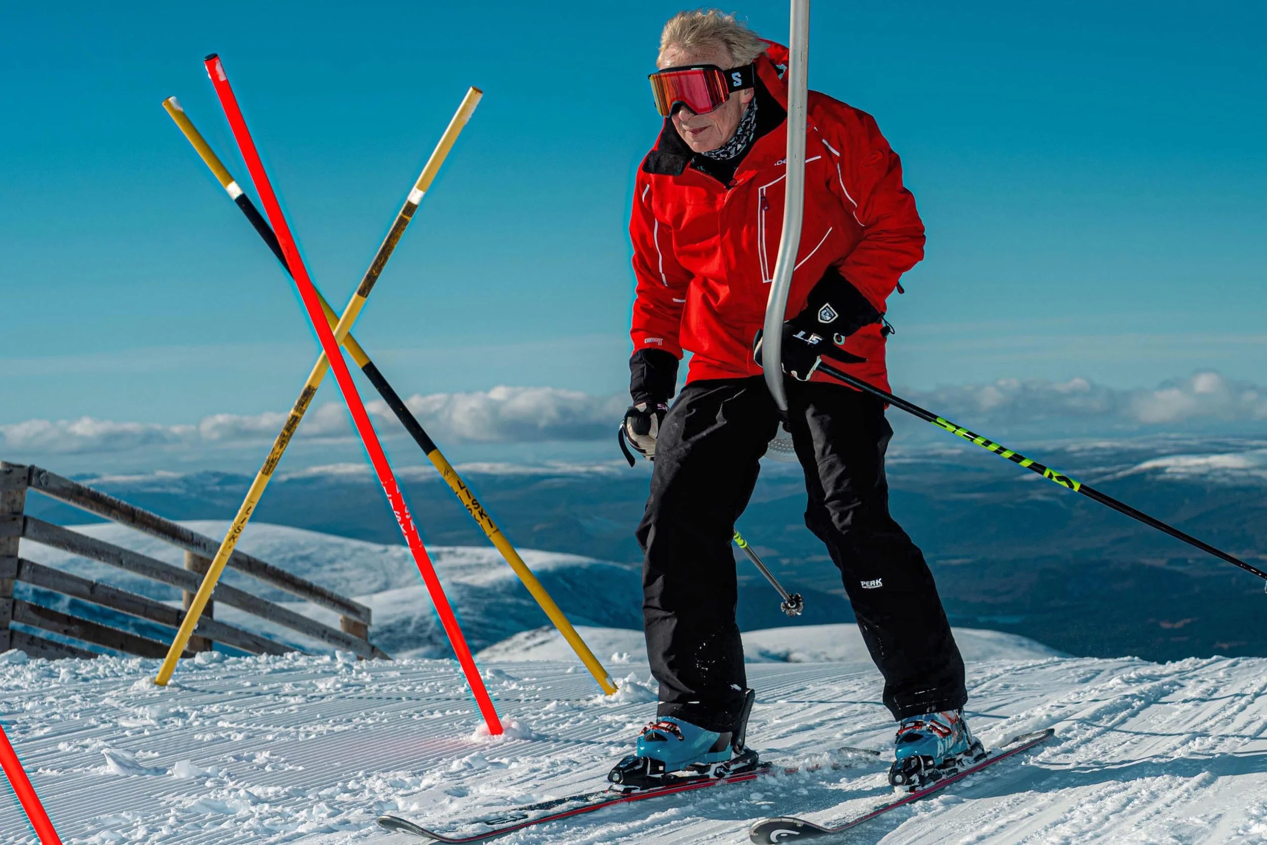 A skier in a red jacket stands beside crossed slalom poles under a blue sky.