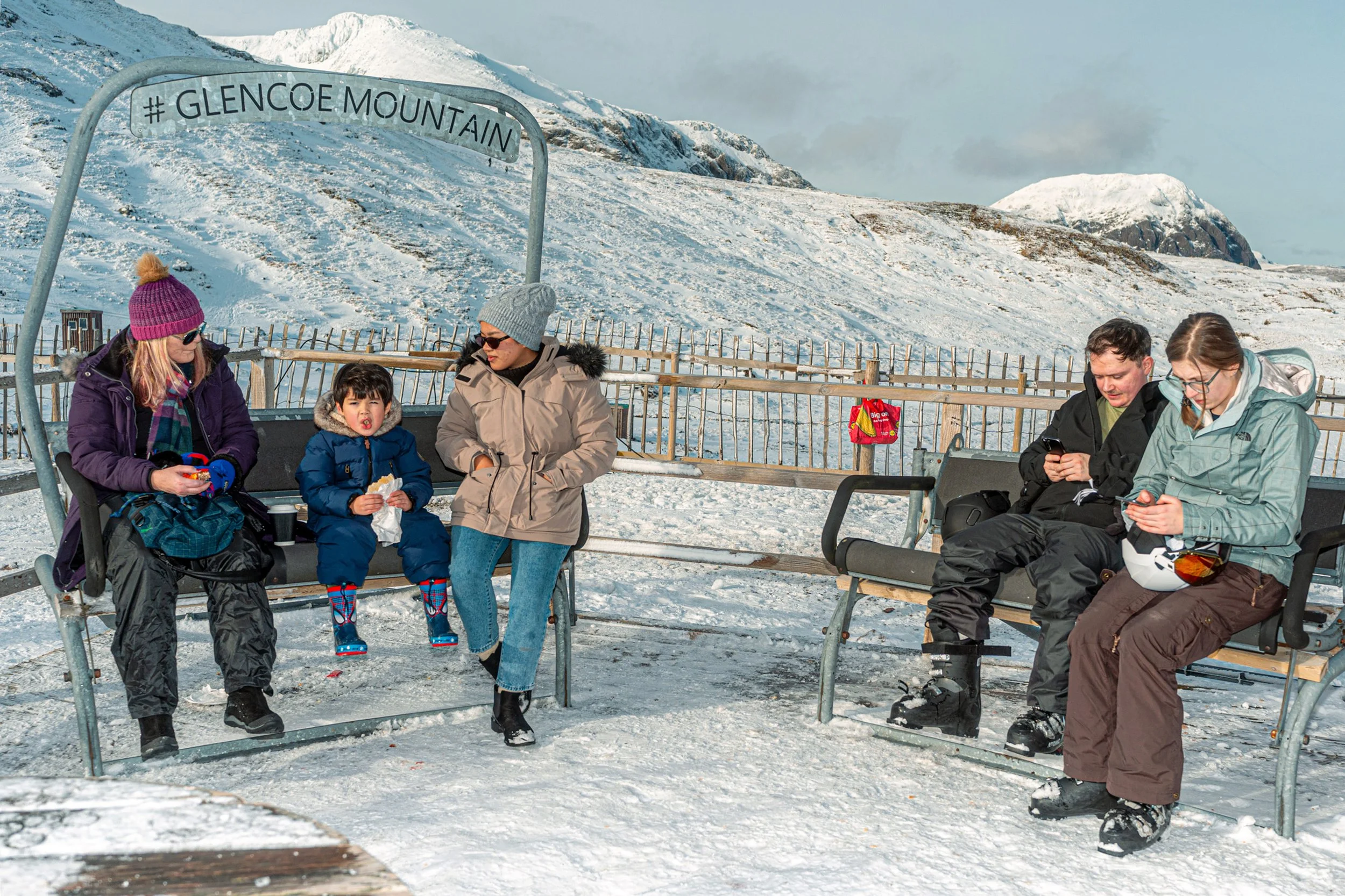 Visitors sit on old chairlift seats marked Glencoe Mountain, with snowy peaks behind.
