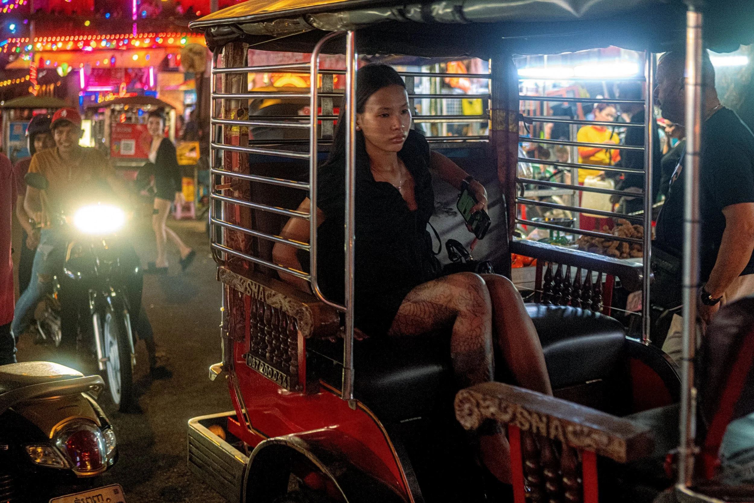 A passenger sits in an open-backed taxi amid neon lights and motorbikes on a busy street.
