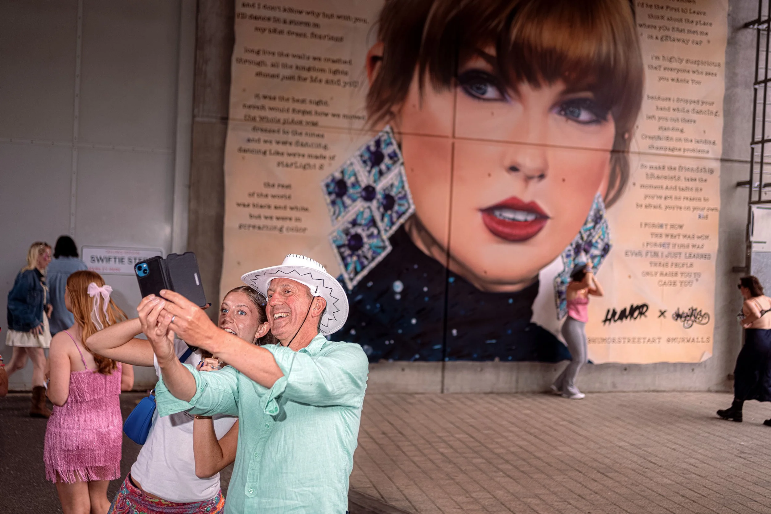 A fan in a turquoise outfit and white cowboy hat takes a selfie in front of a giant Taylor Swift poster.