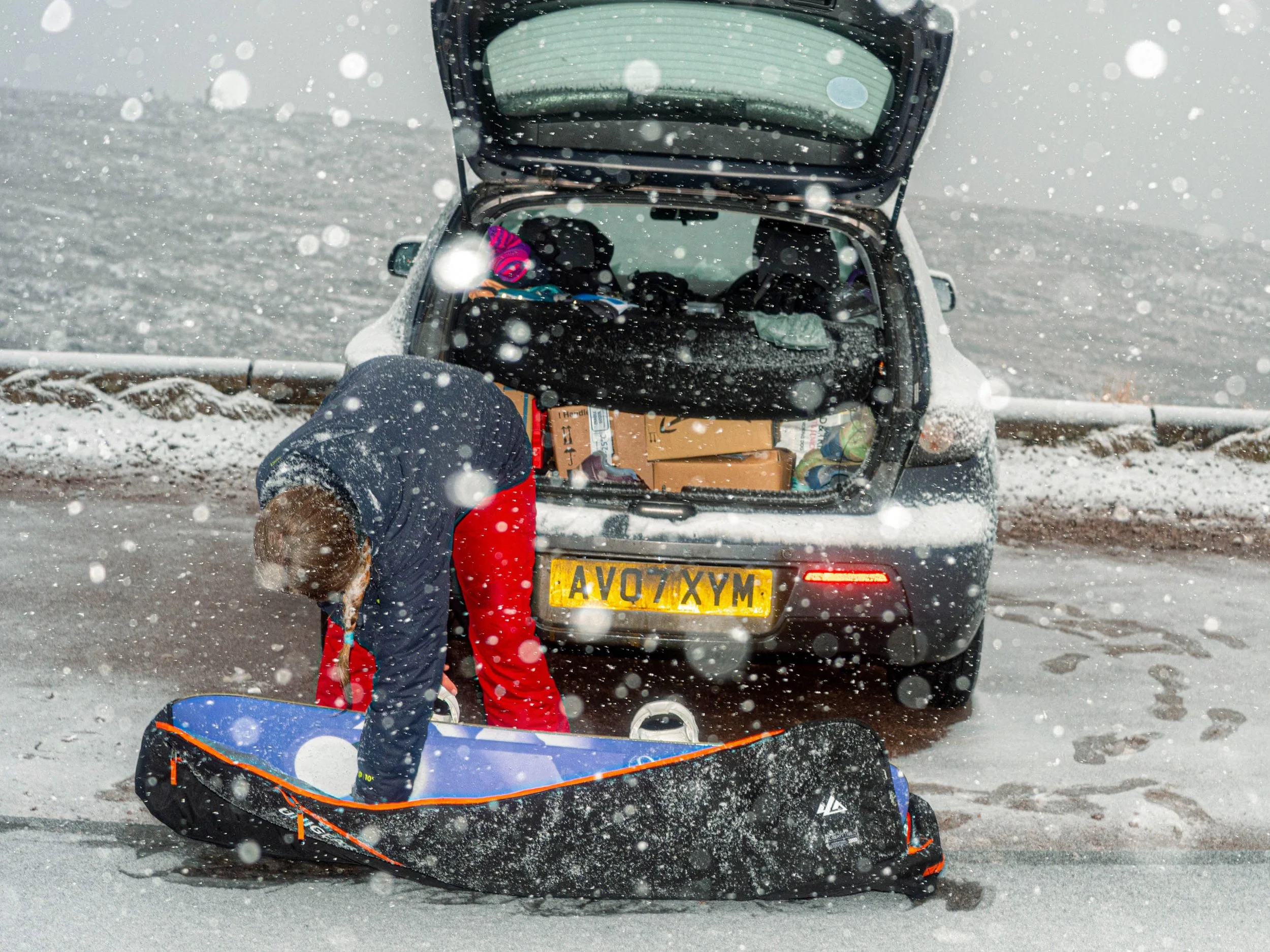 A man packs a board bag beside his car boot as snow falls in the car park.