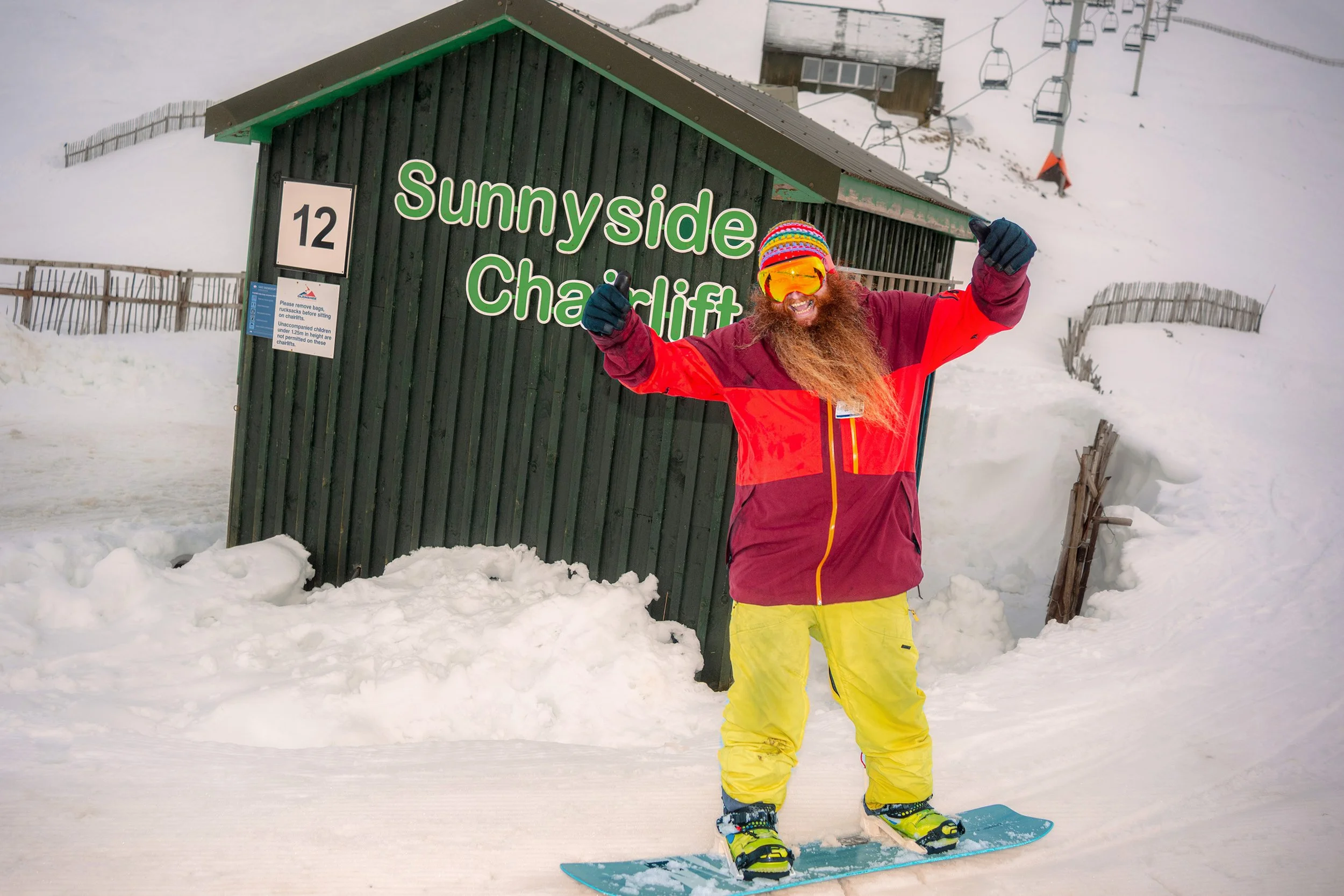 A bearded snowboarder in a red jacket and yellow trousers poses with arms raised beside the Sunnyside Chairlift hut.