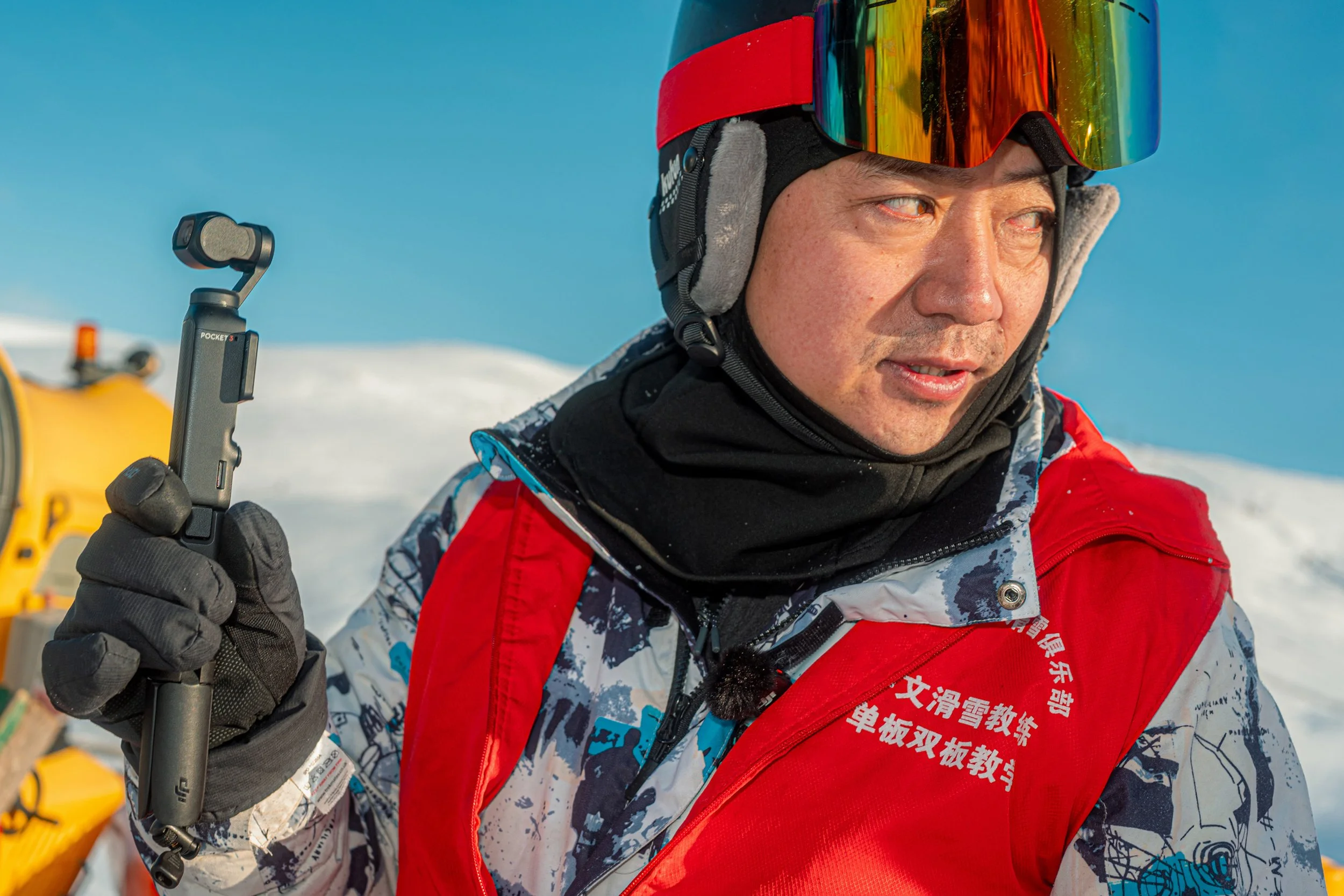A skier in mirrored goggles and a red bib with Chinese lettering holds a handheld camera gimbal.
