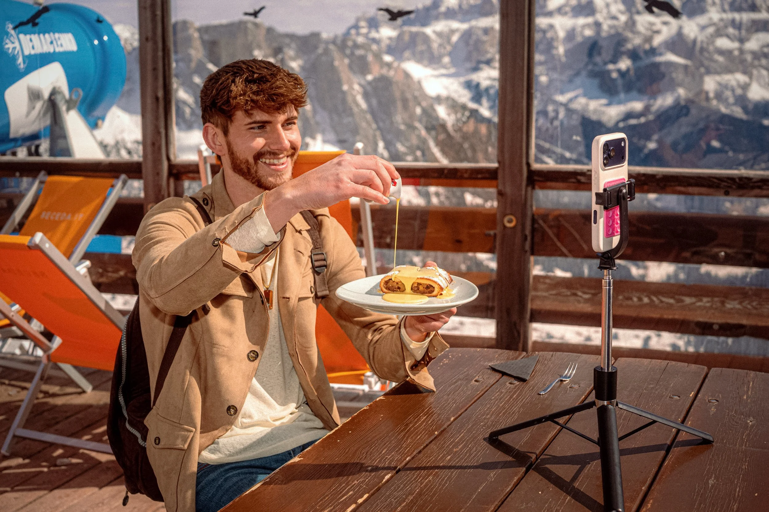 A smiling diner lifts food towards a phone on a tripod at an outdoor table overlooking snowy mountains.