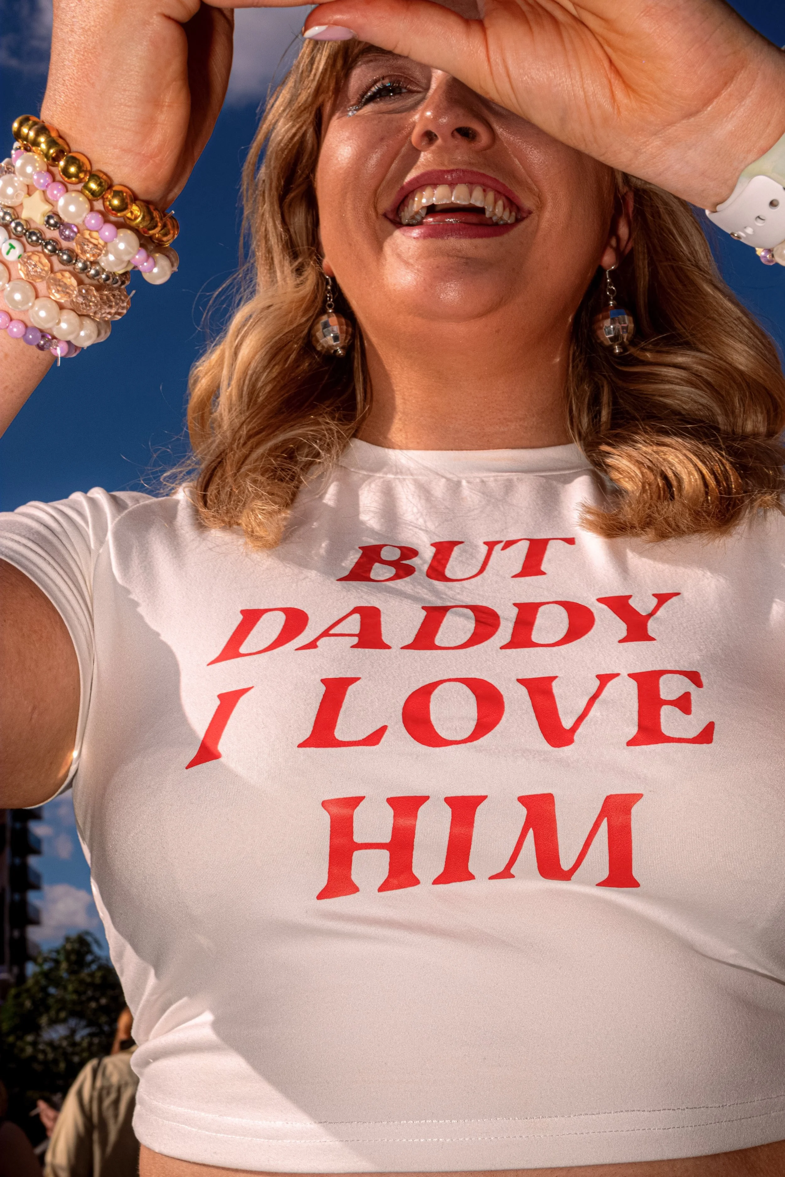 A laughing Taylor Swift fan raises her arms while wearing a white T-shirt reading “But Daddy I Love Him”.