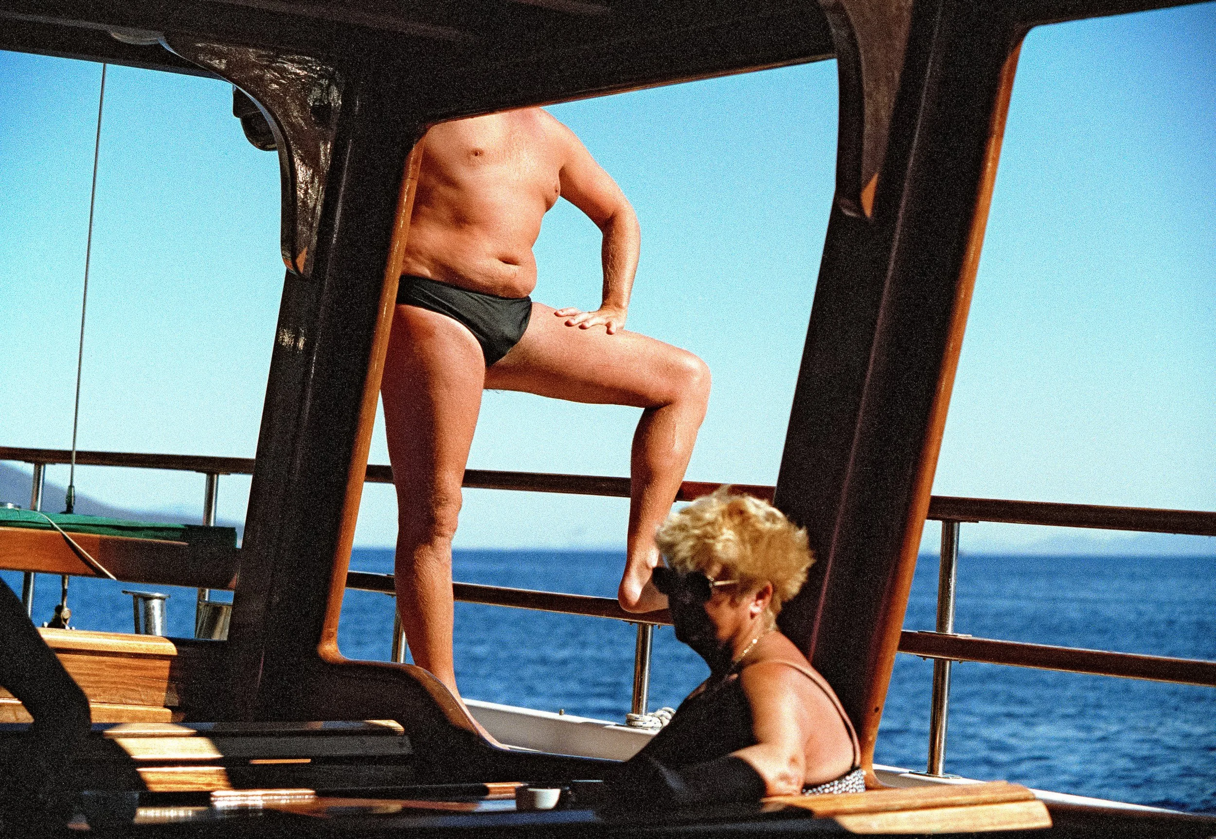 Holidaymakers in swimwear stand and sit among wooden beams on a boat at sea.