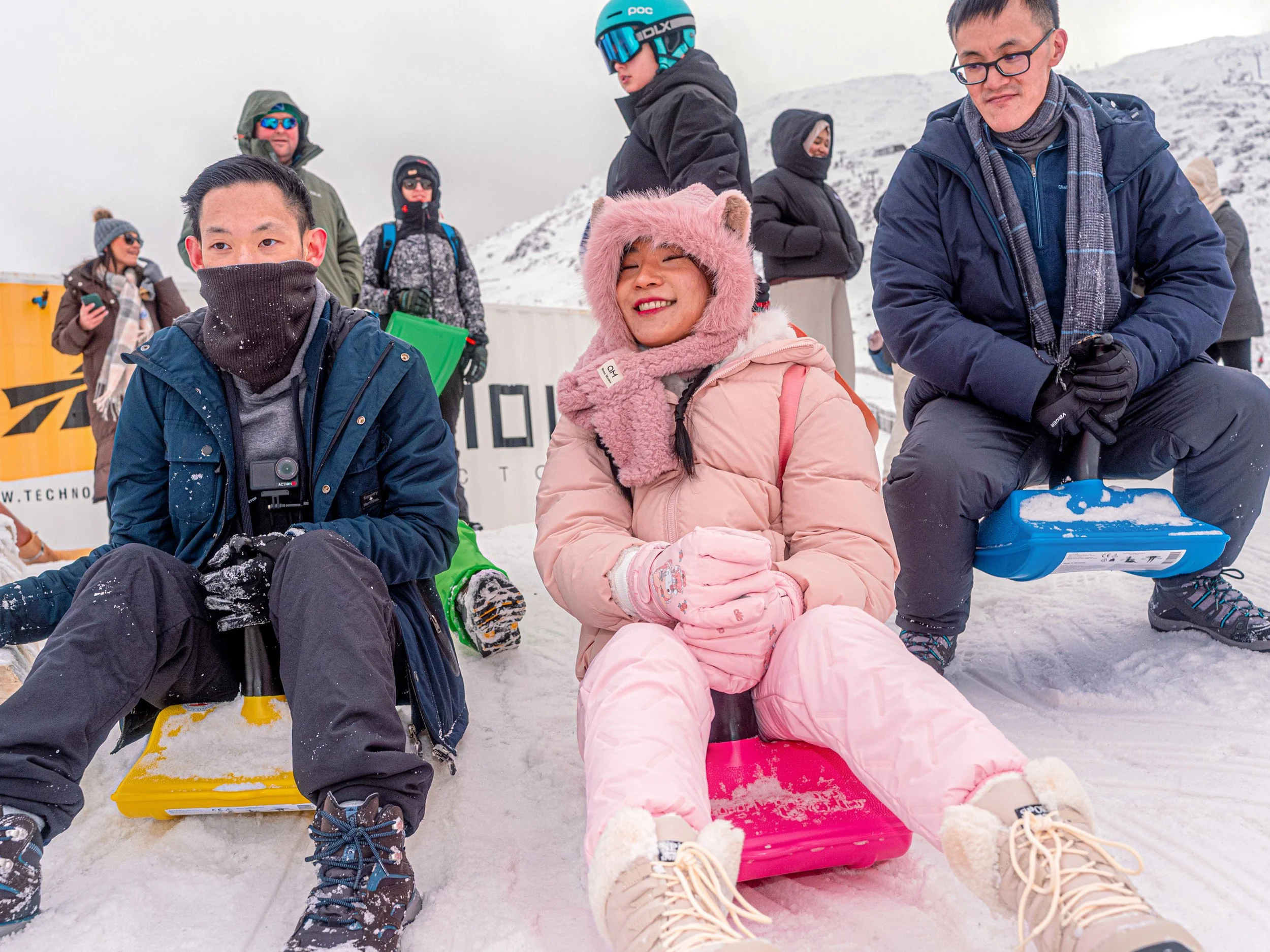 Visitors in winter coats sit on plastic sledges in the snow, one in a pink furry hat.