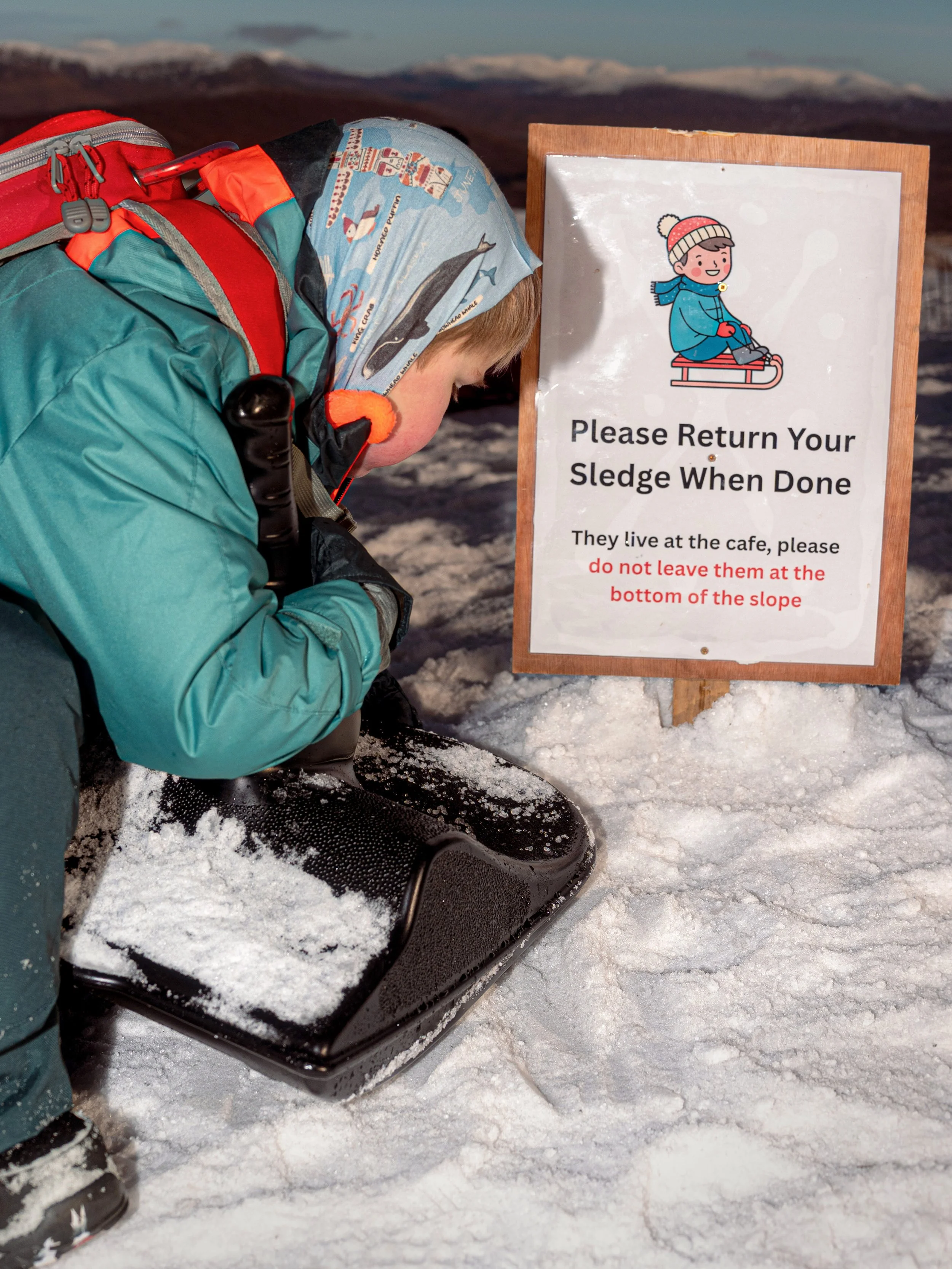 A child in a teal jacket and headscarf picks up a sledge beside a sign asking visitors to return their sledges.