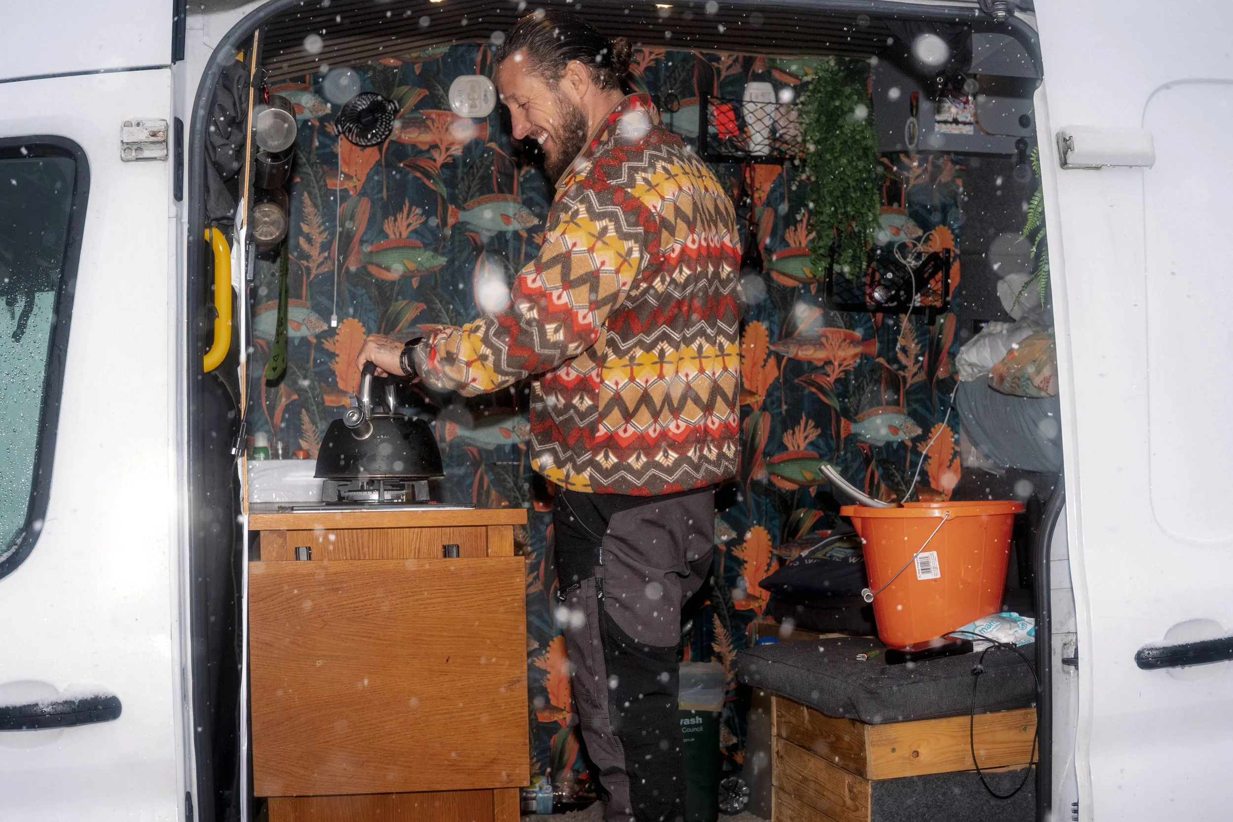 A man in a patterned jumper makes a hot drink in the back of his camper van as snow falls.