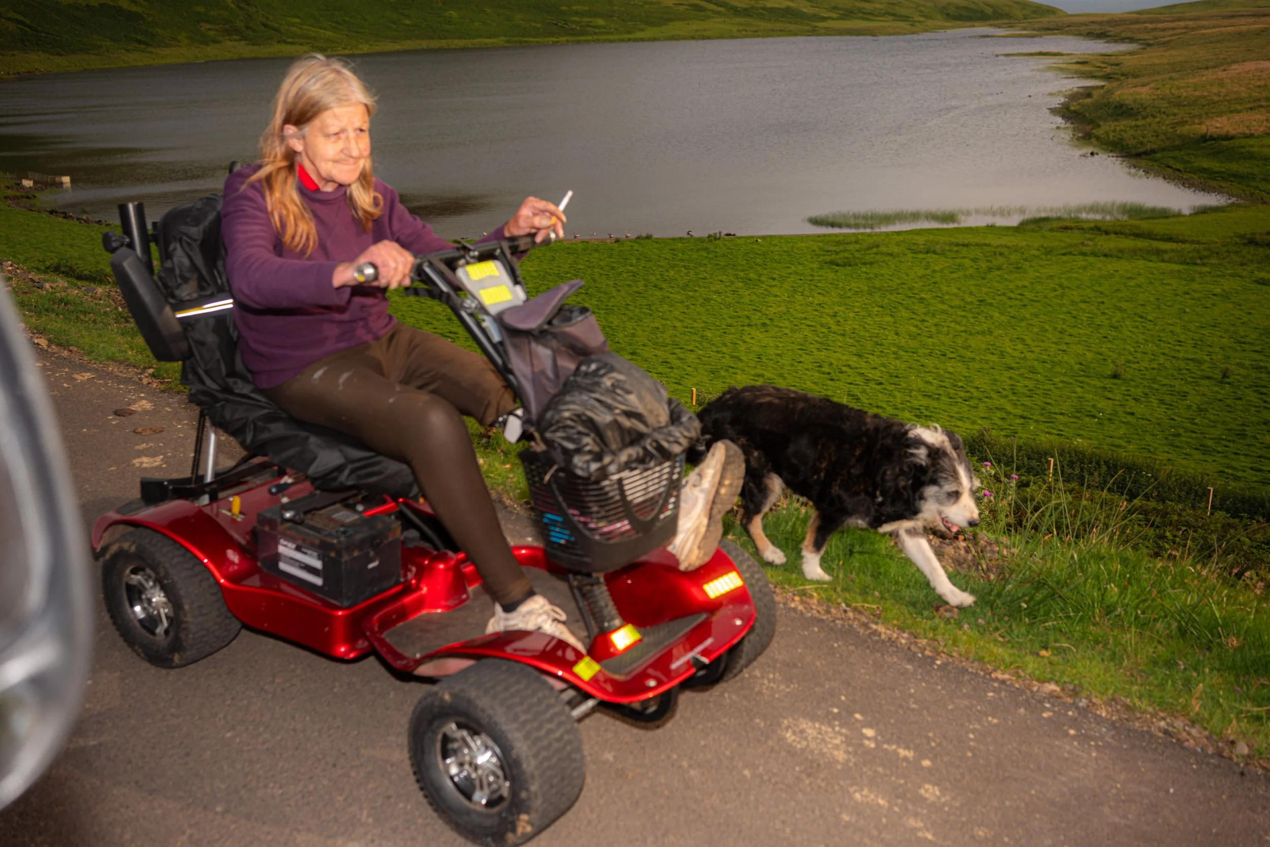 A woman rides a red mobility scooter along a loch-side path with a border collie running beside her.