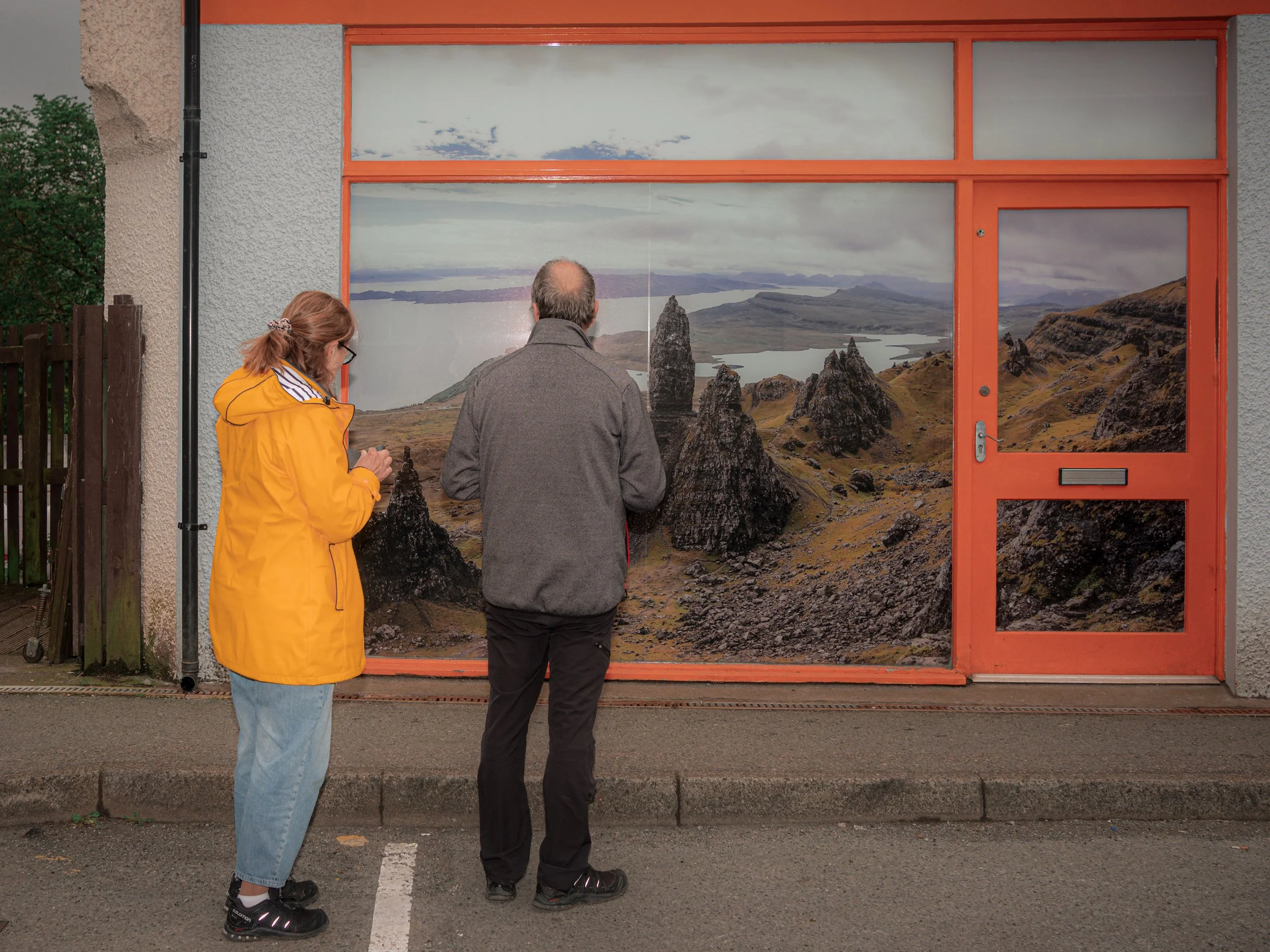 Two people stand in front of a shop window covered with a picture of a rocky Skye landscape.