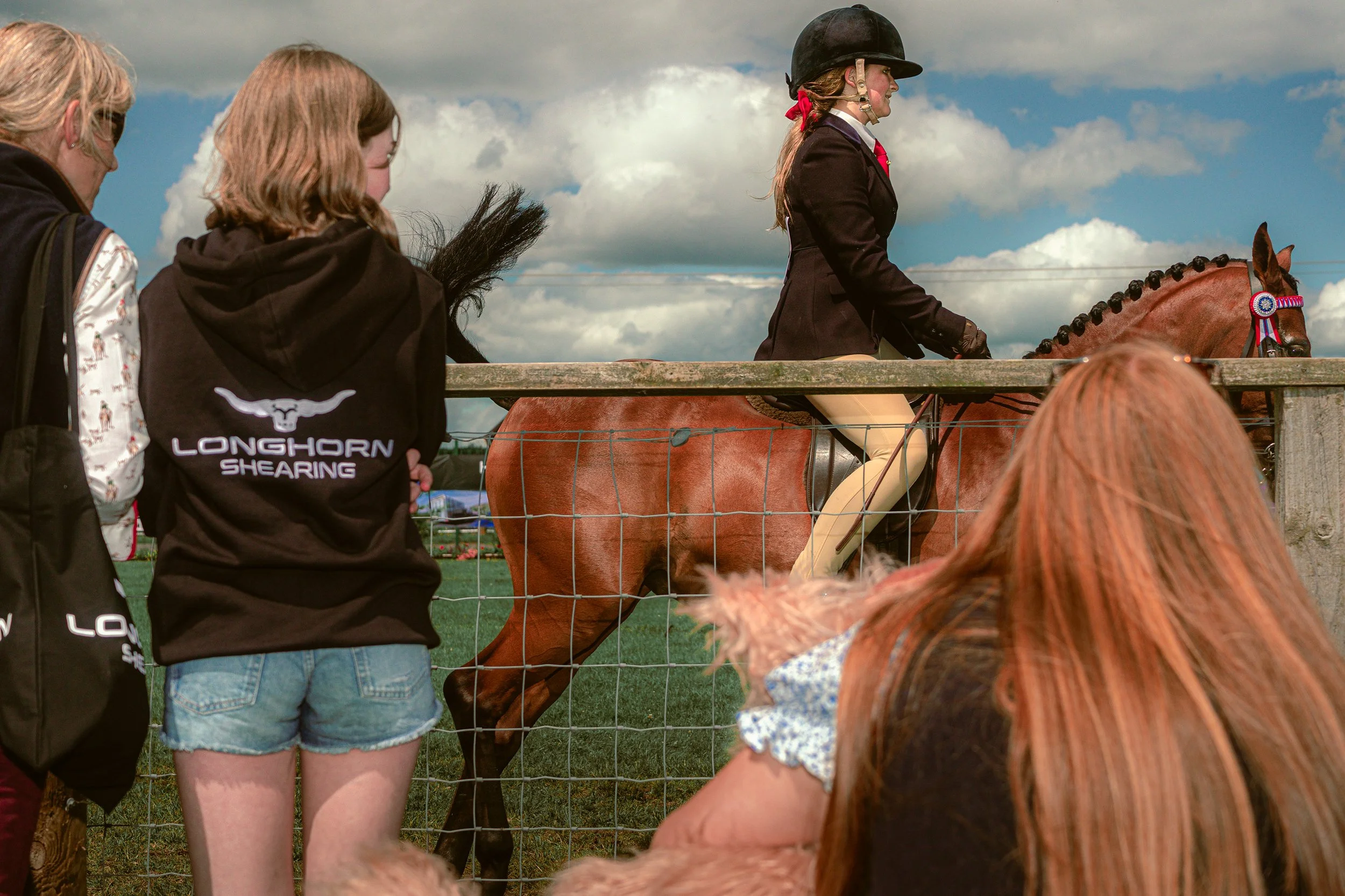 Spectators lean on a fence to watch a young rider on a horse at an agricultural show.