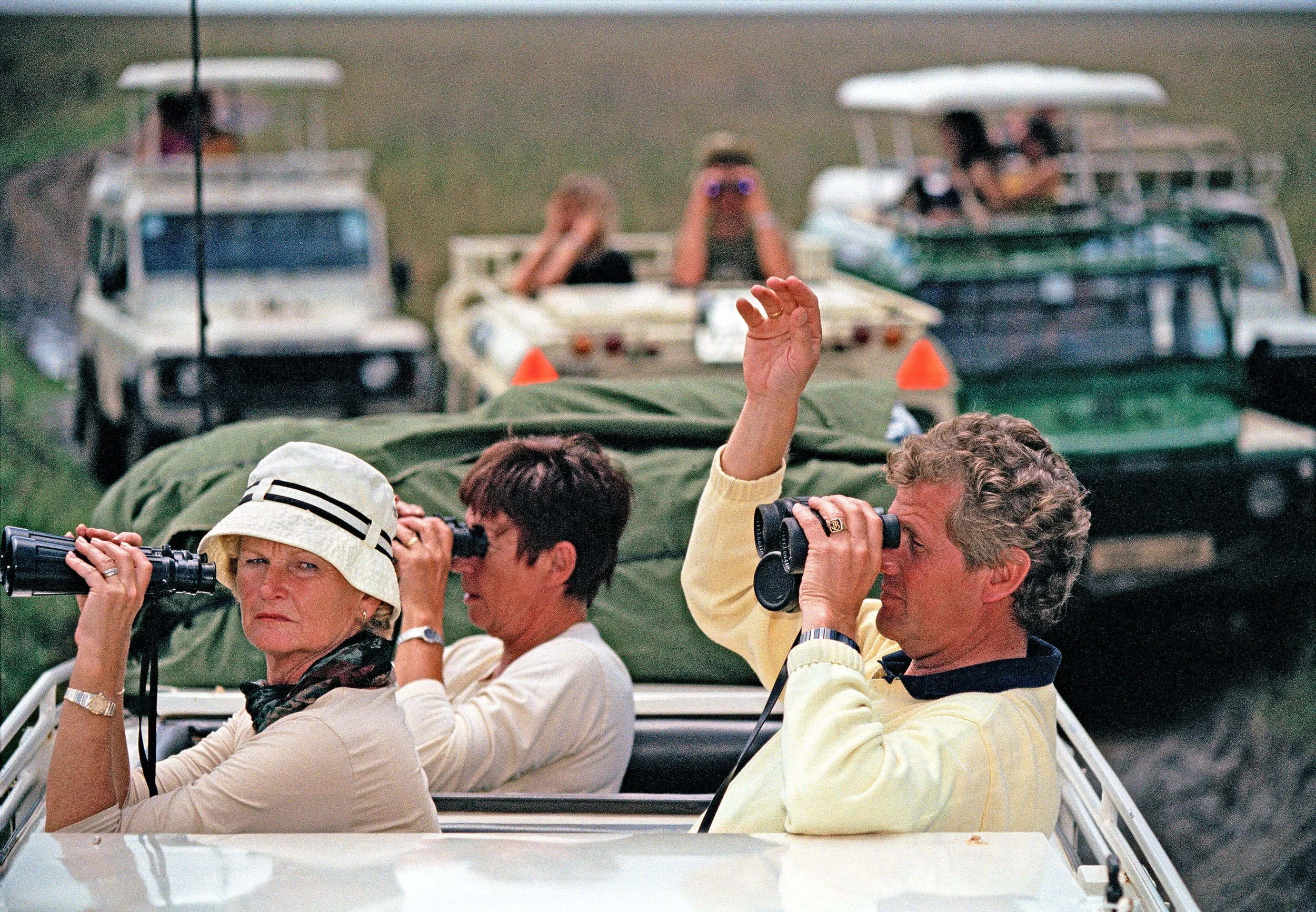Tourists raise cameras and binoculars from open safari vehicles on grassland.