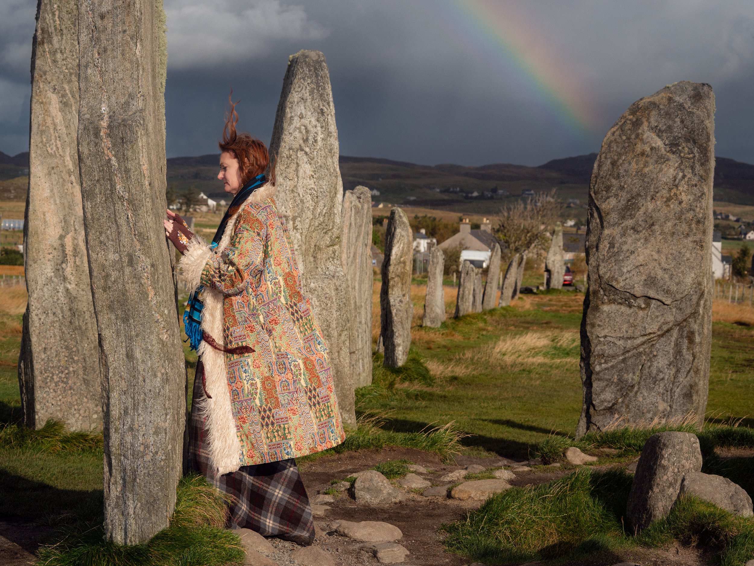 A woman in a patchwork coat stands among standing stones under a rainbow.