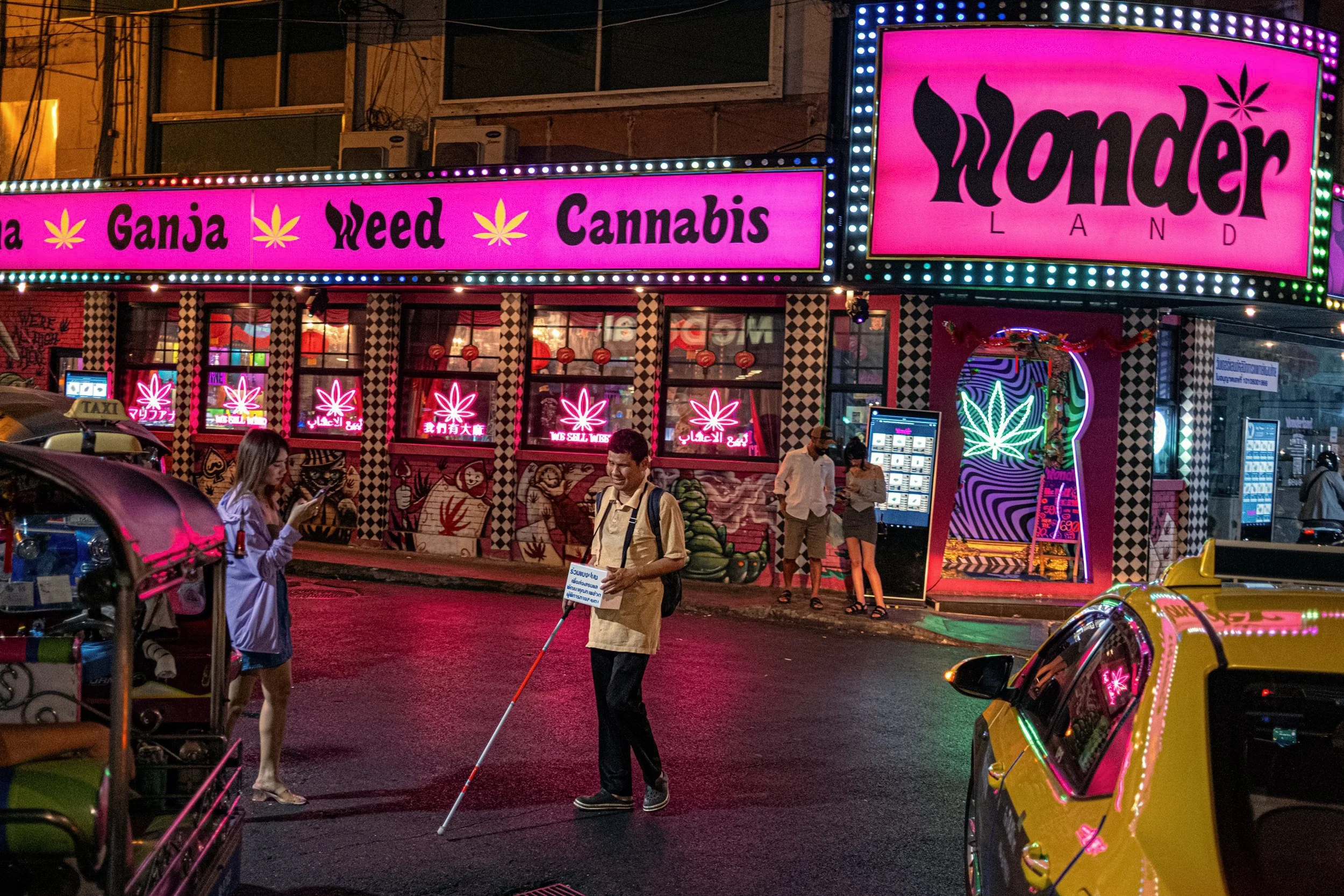 A neon-lit street in Thailand with a cannabis shop, pedestrians and taxis.