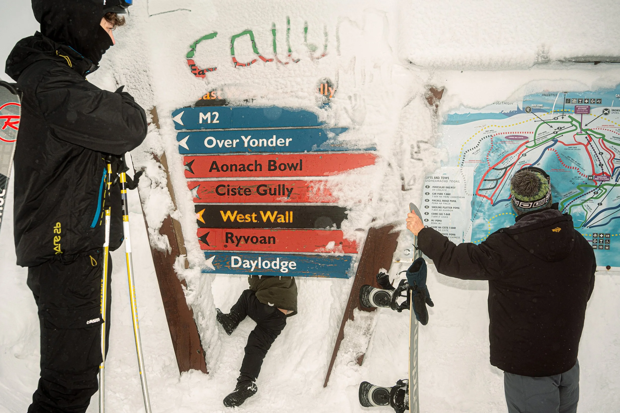 Two snowboarders stand beside a snowy signpost pointing to runs including Aonach Bowl, West Wall and Ryvoan.