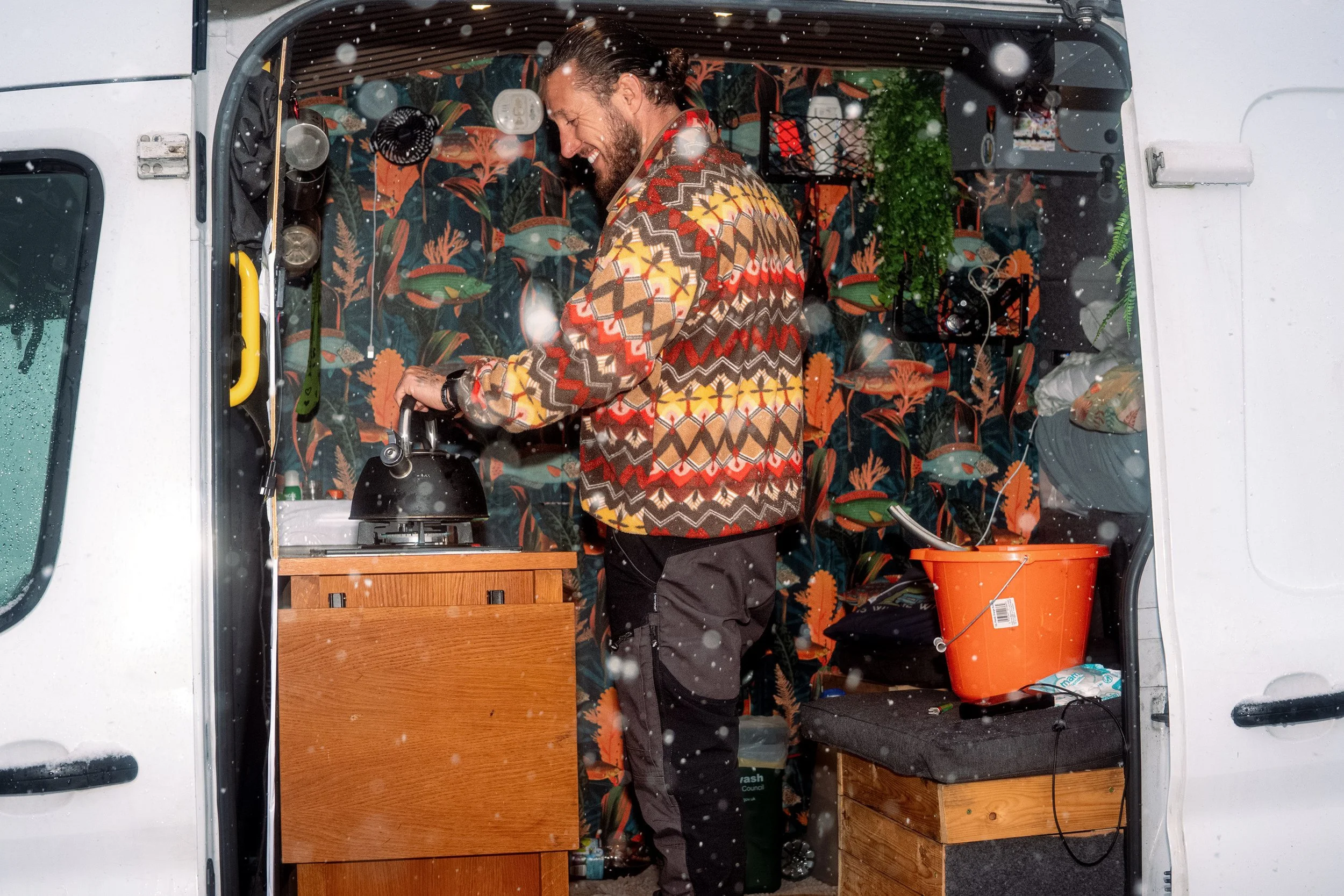 A person in a patterned jumper prepares a drink inside a campervan during snowfall, from Weather Permitting.
